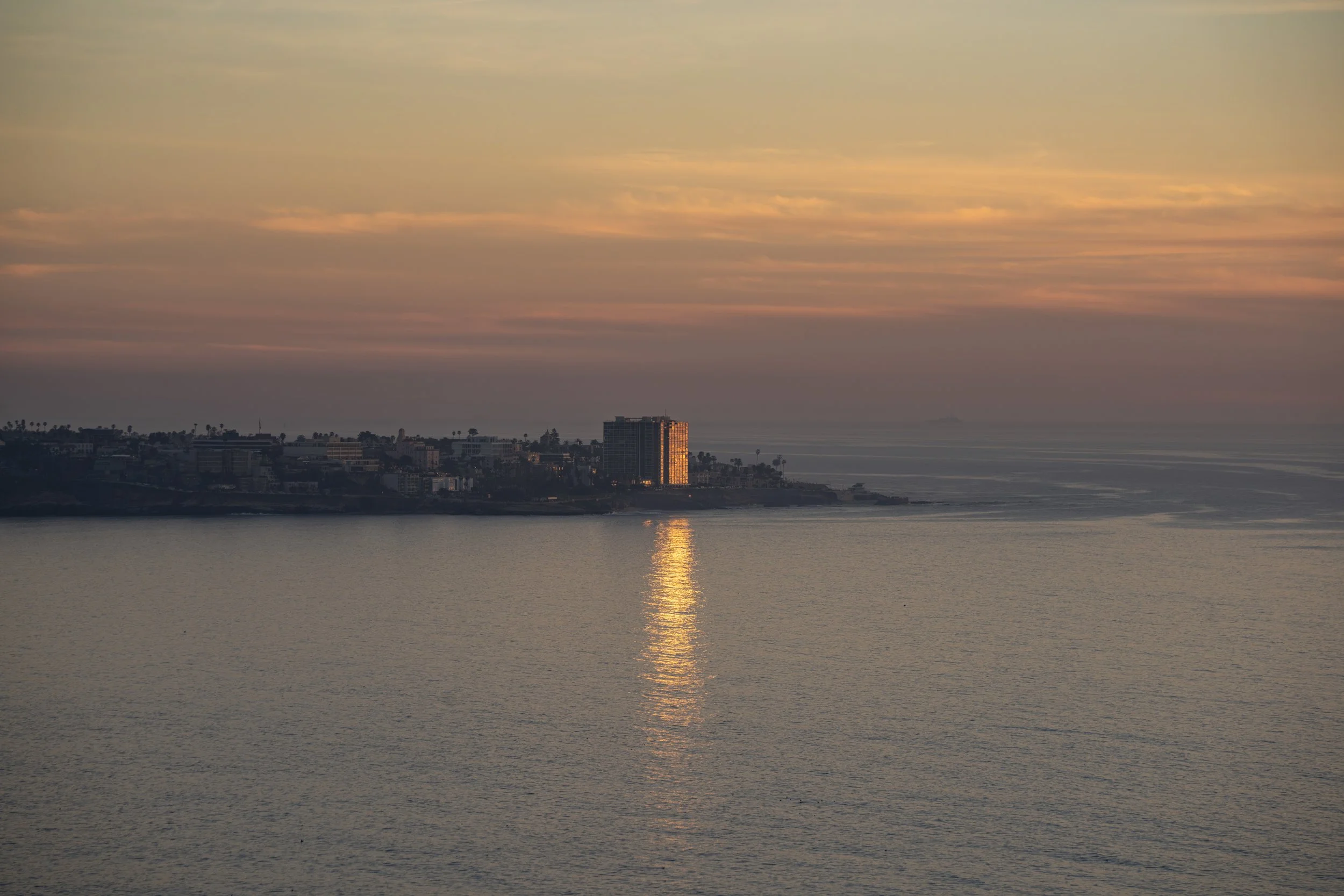 Sunset over the ocean with reflections on the water, and a distant cityscape with buildings and palm trees on the horizon.