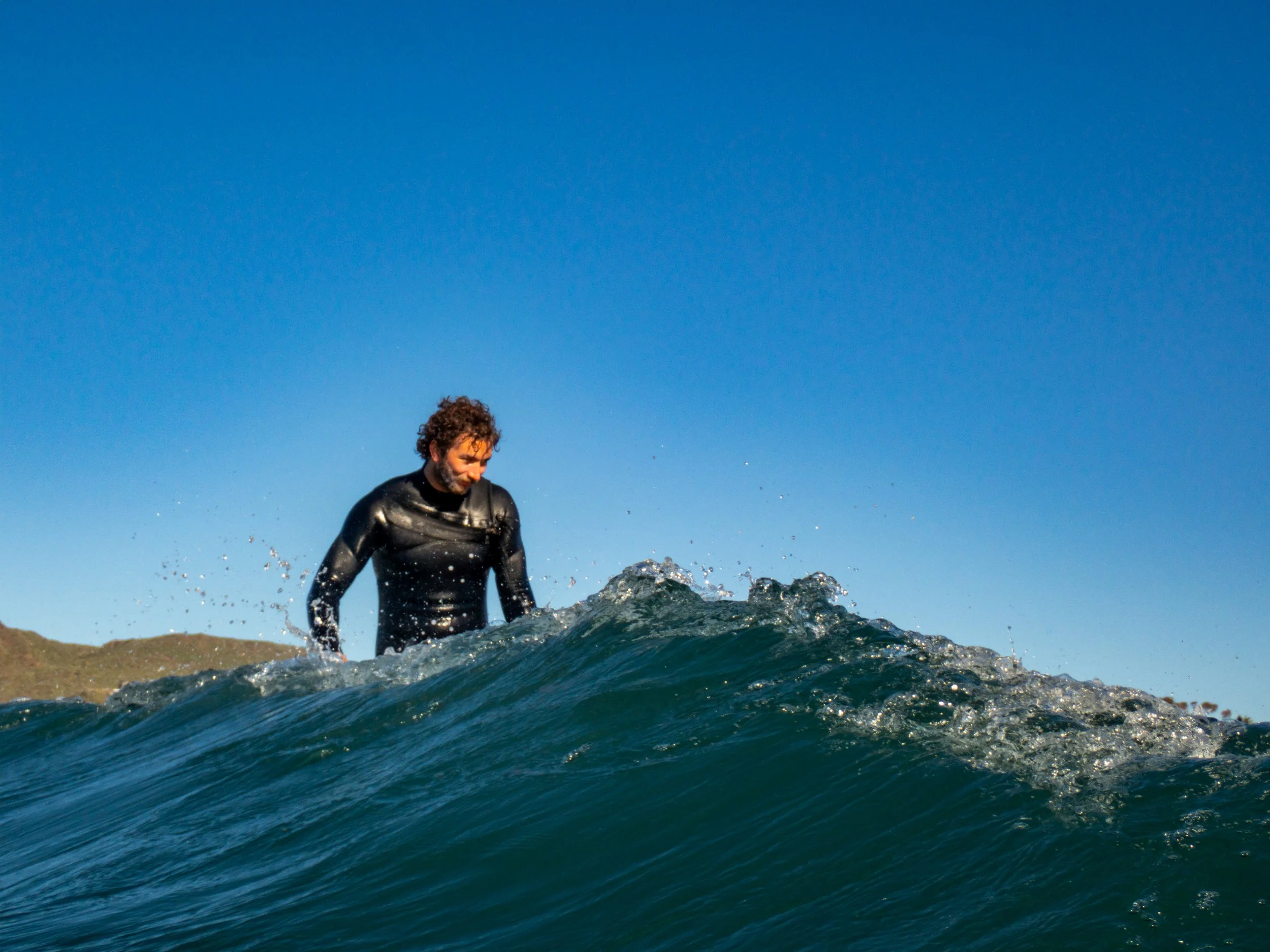 Man in a black wetsuit standing on a surfboard on a wave with a clear blue sky in the background.