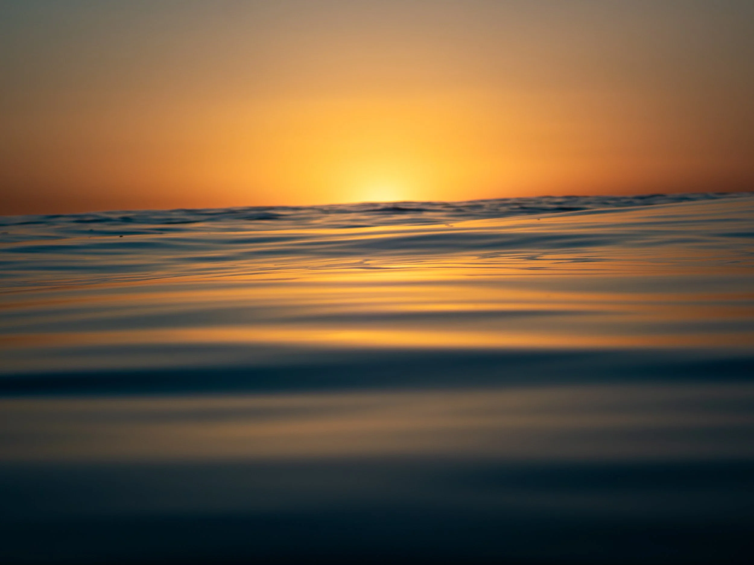 Calm ocean water with gentle ripples during sunset or sunrise, with the sun partially visible on the horizon and a colorful sky transitioning from dark to bright.