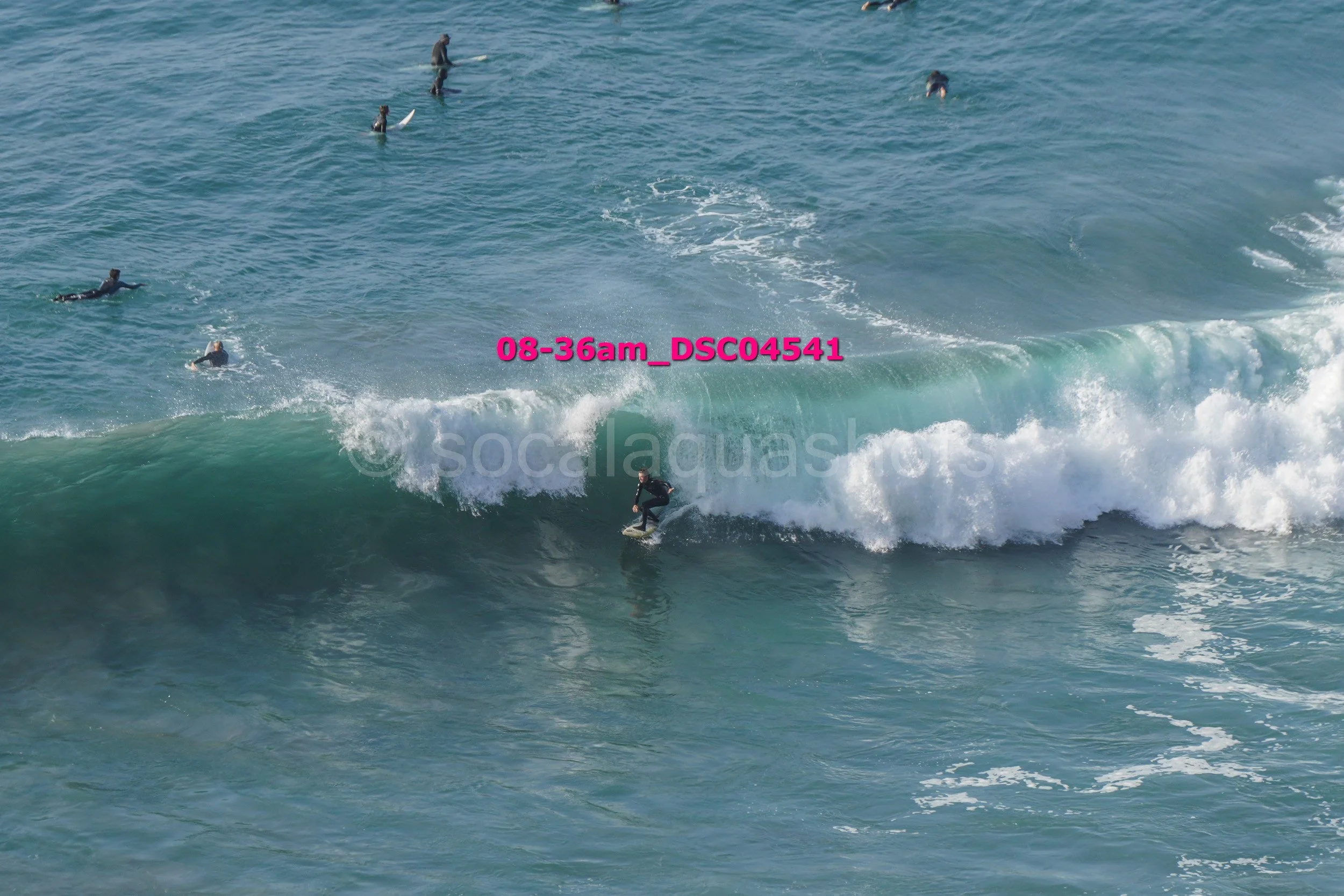 Surfer riding a wave near shore with several people in wetsuits in the water surrounding him.