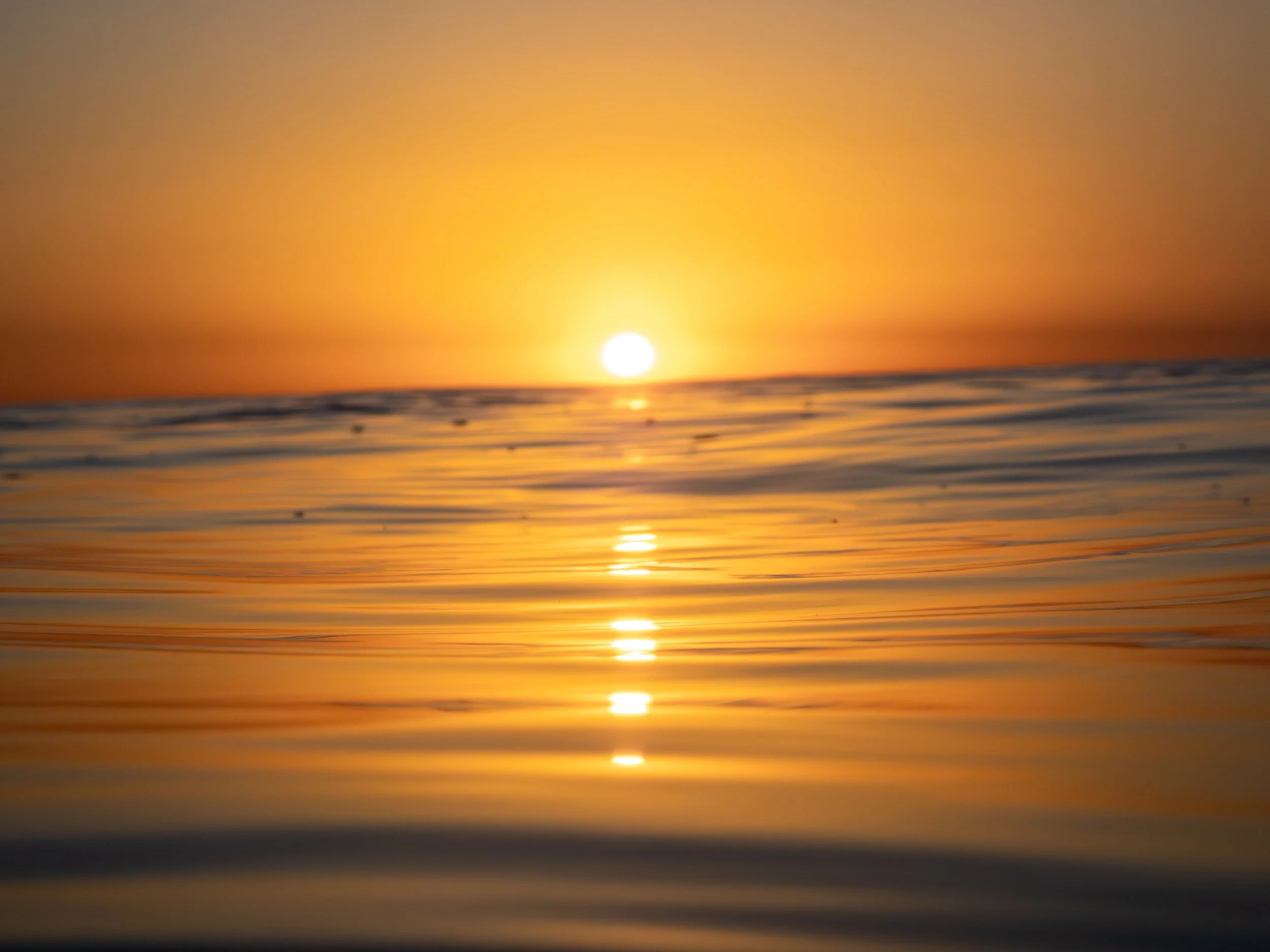 Sunset over calm ocean water with the sun low on the horizon and its reflection visible on the water's surface.