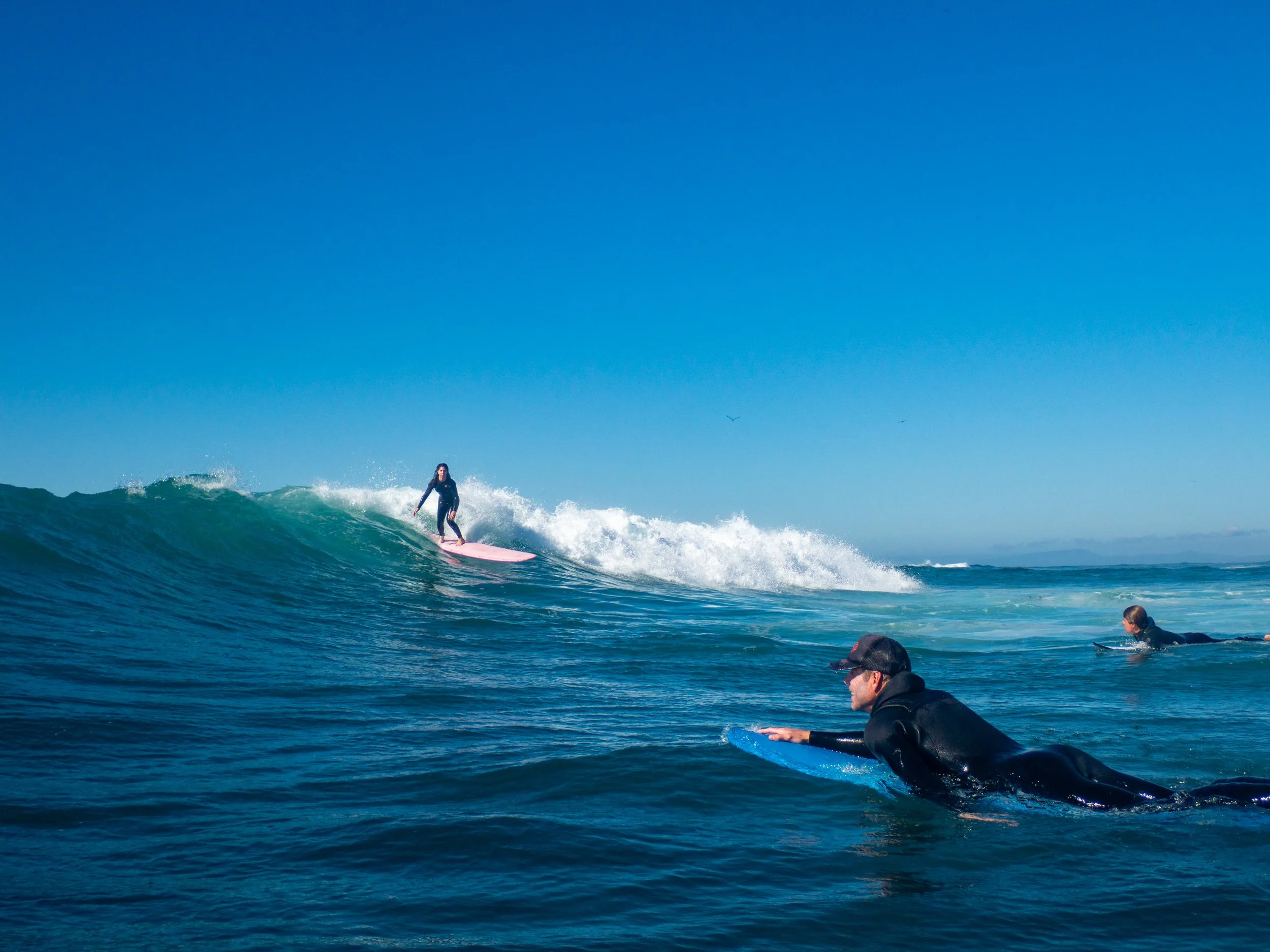 A group of surfers, including a woman, is surfing on the ocean waves under a clear blue sky.