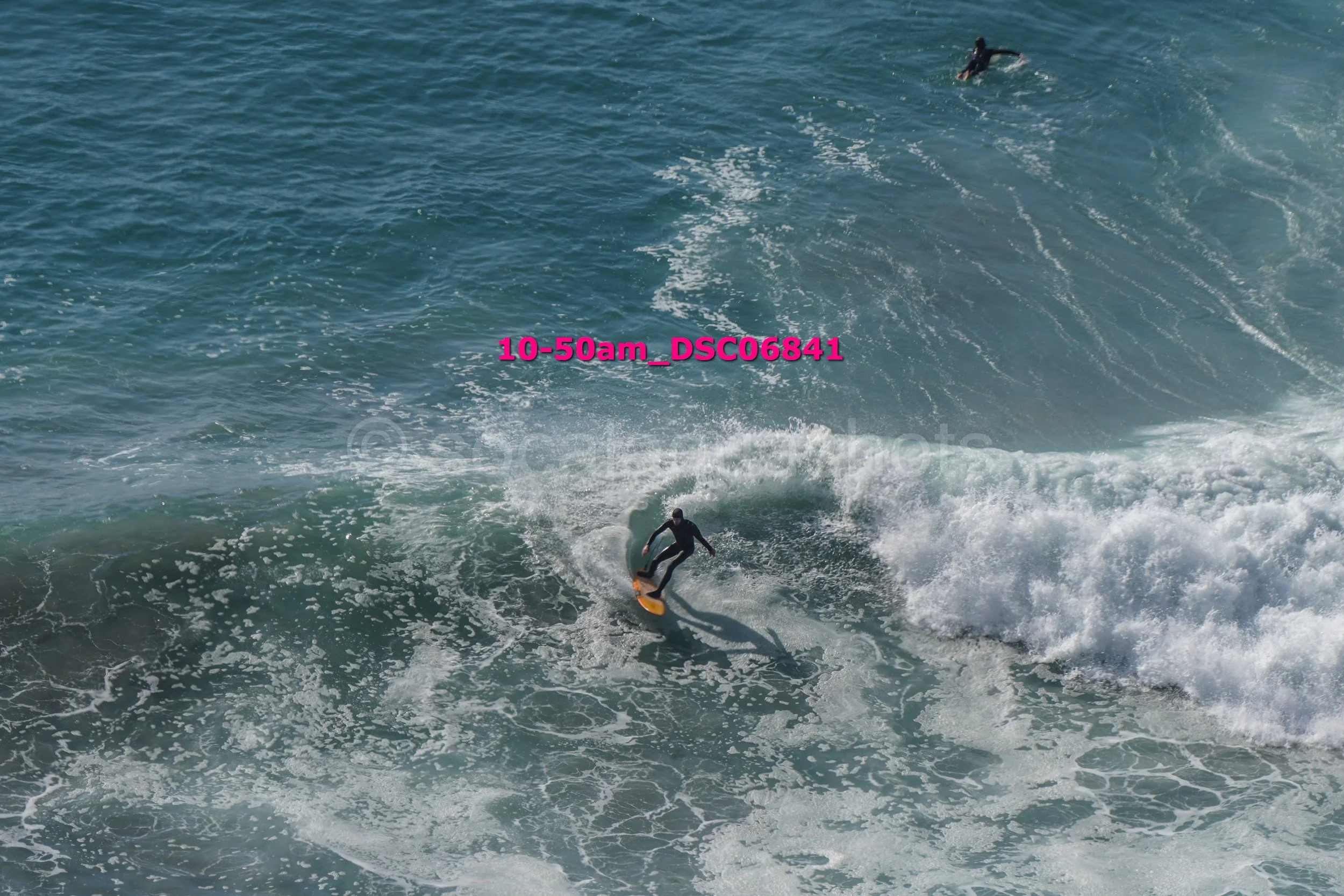 A person wearing a wetsuit surfing on a wave with another surfer in the background in the ocean.