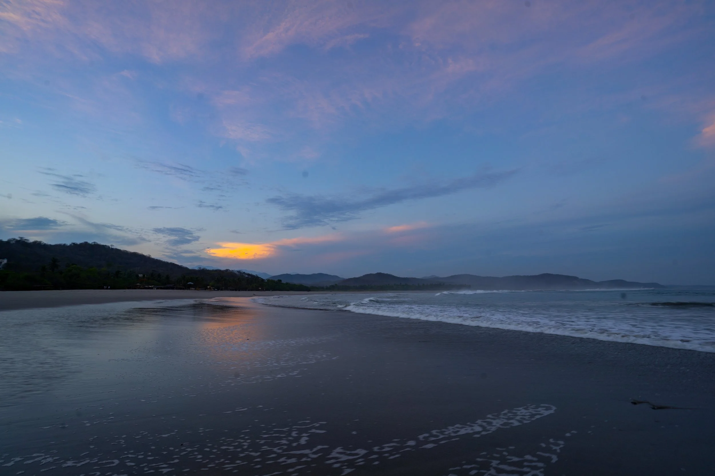 Sunset over a beach with gentle waves and a mountain range in the background, colors shifting from yellow to purple in the sky.