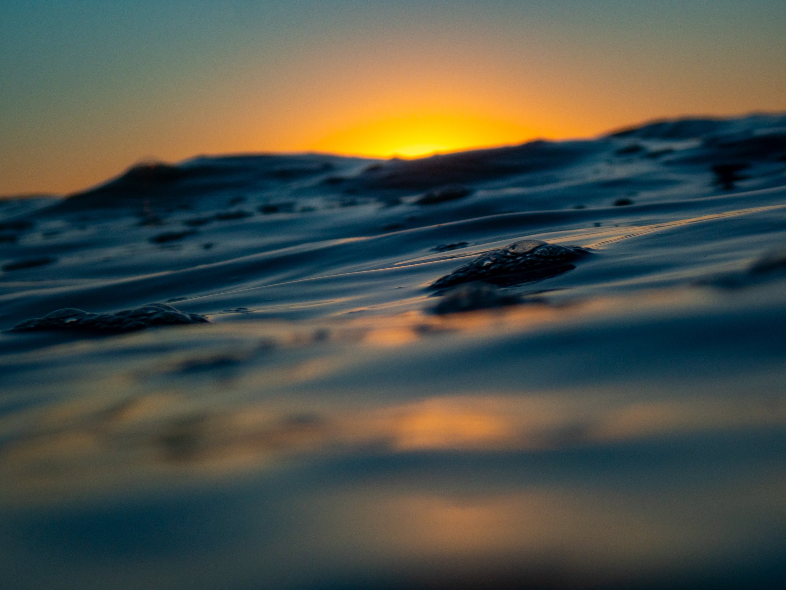 A close-up view of ocean water during sunset, with gentle waves and the sun partially visible on the horizon.