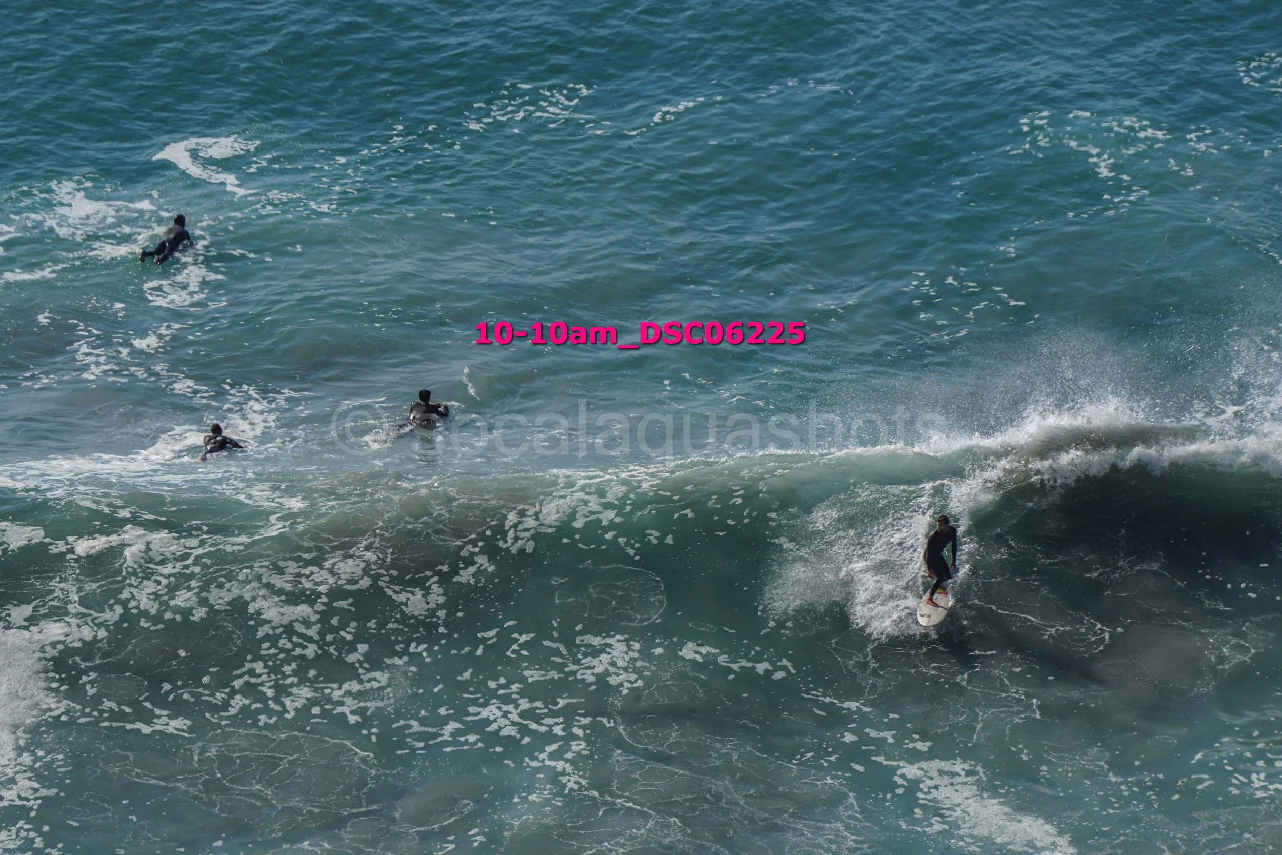Surfer riding a wave with three other surfers in the ocean and a group of people swimming nearby.