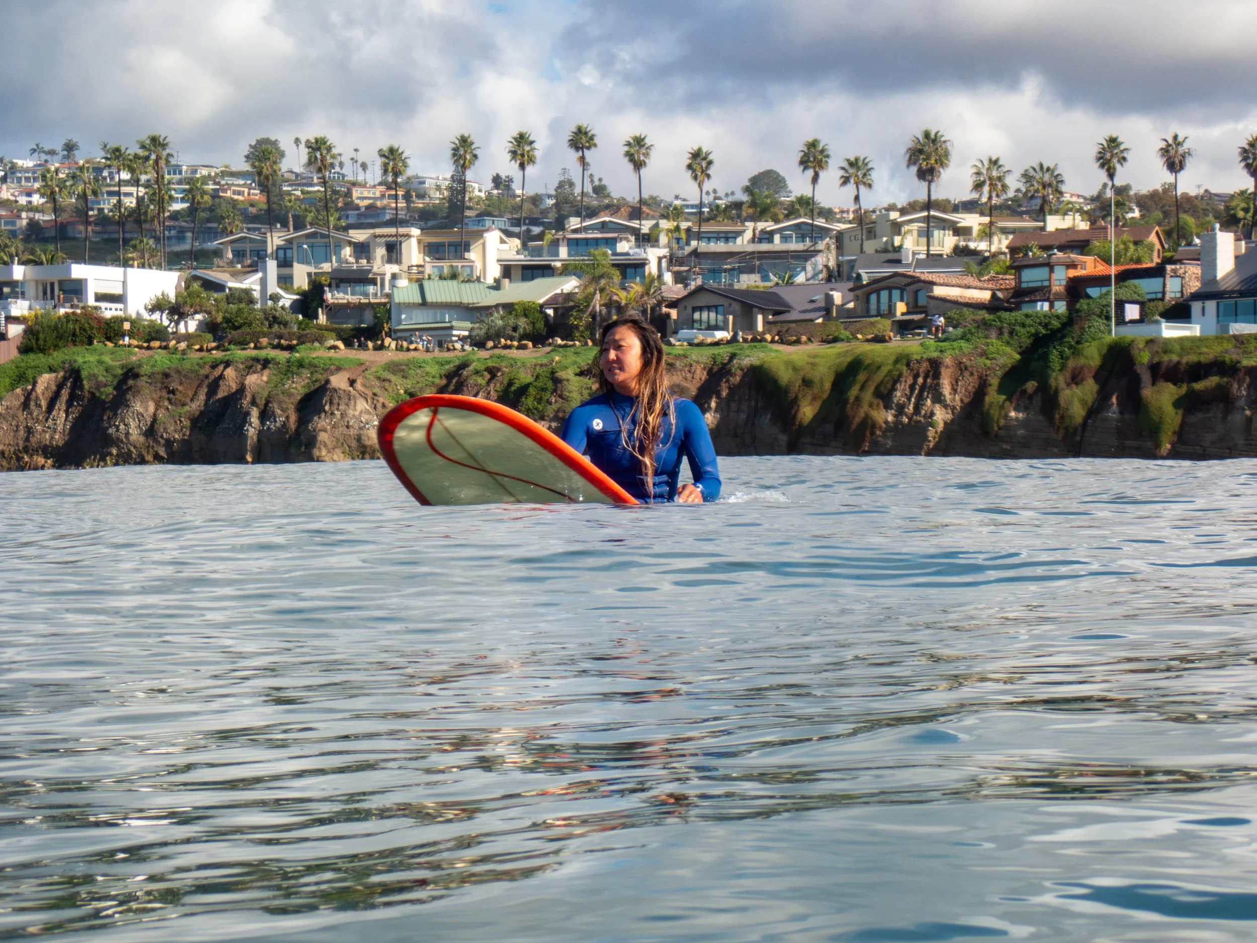 Woman in a blue wetsuit holding a surfboard in the ocean near a coastal residential area with palm trees and houses.