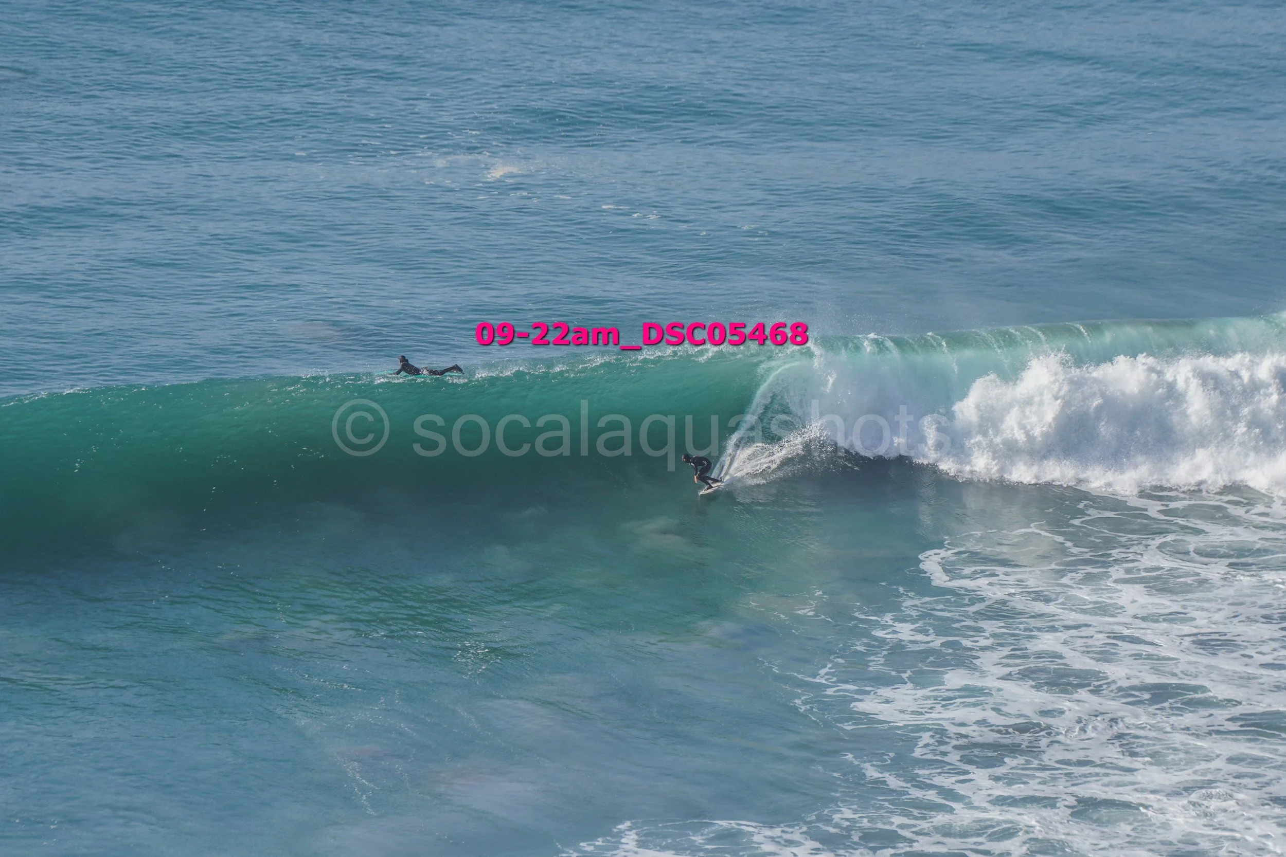 A surfer riding a large wave in the ocean with another surfer floating on the water nearby.