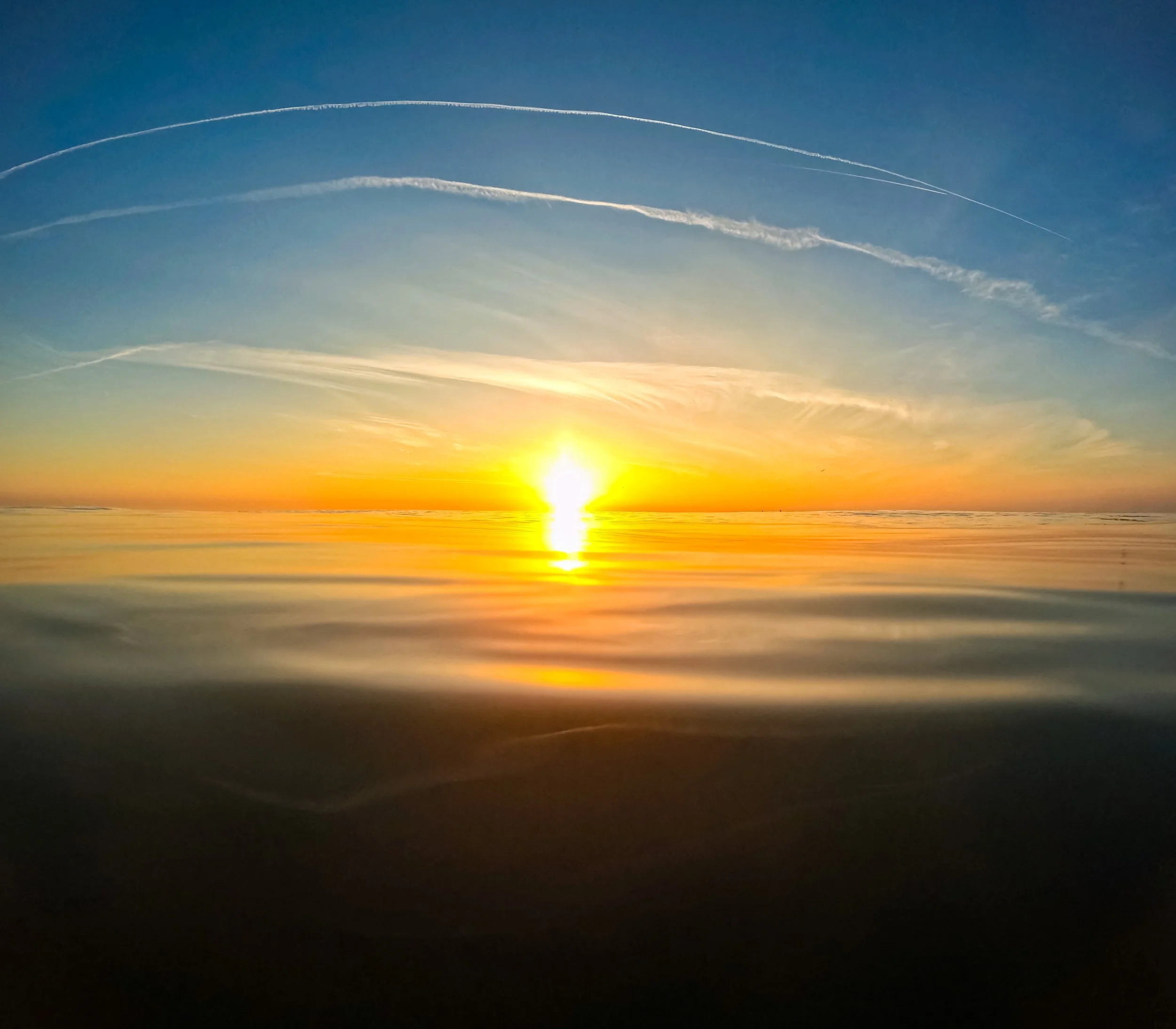 Sunset over the ocean with a clear sky and a few clouds, showing streaks of plane contrails.