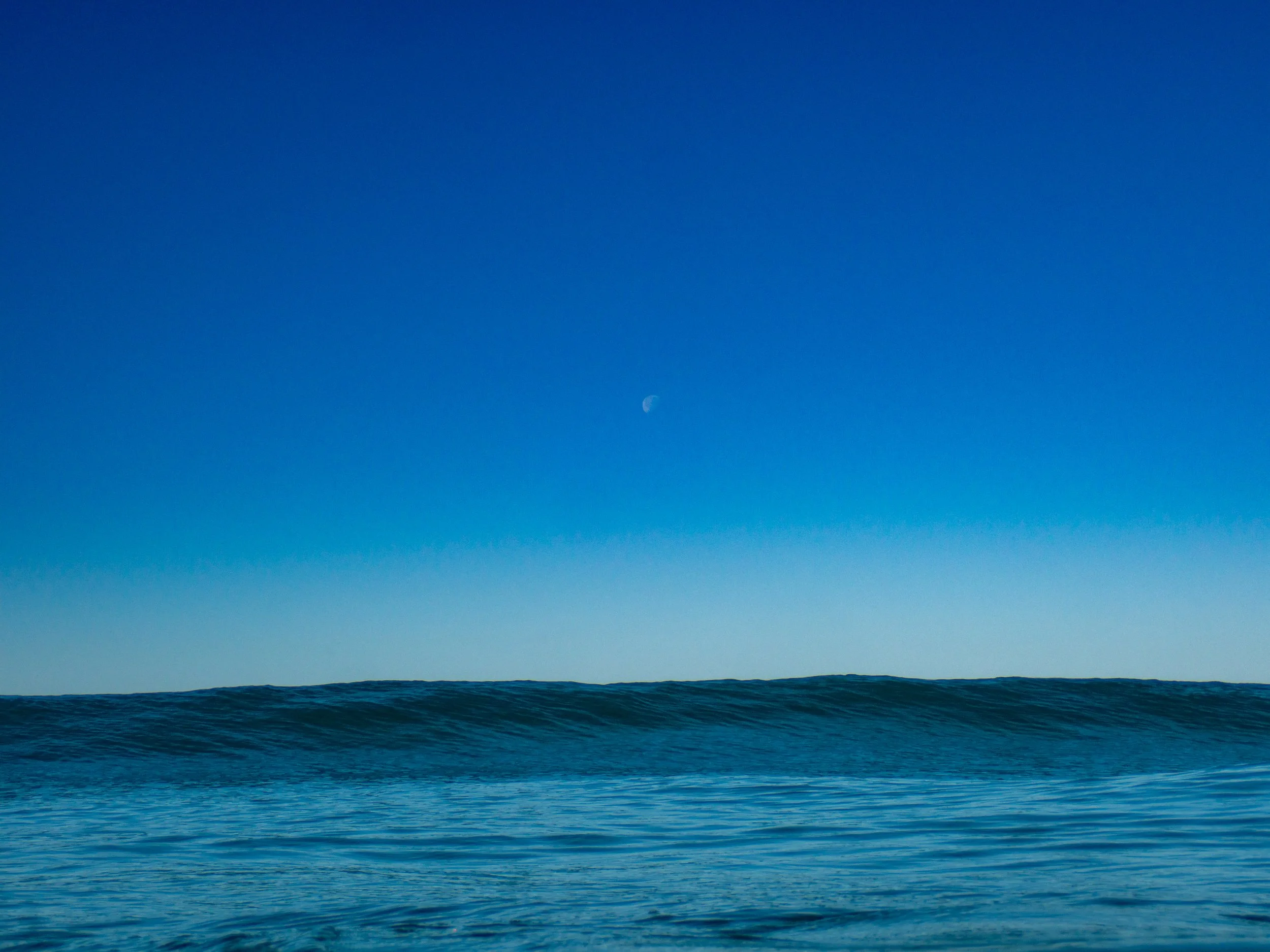 Ocean waves under a clear blue sky with the moon visible.