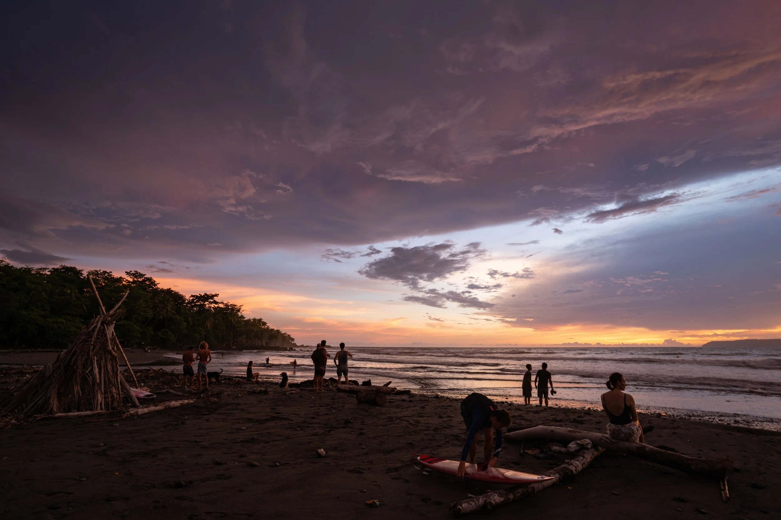People on a beach during sunset, with a colorful sky featuring clouds and pink, purple, and orange hues.