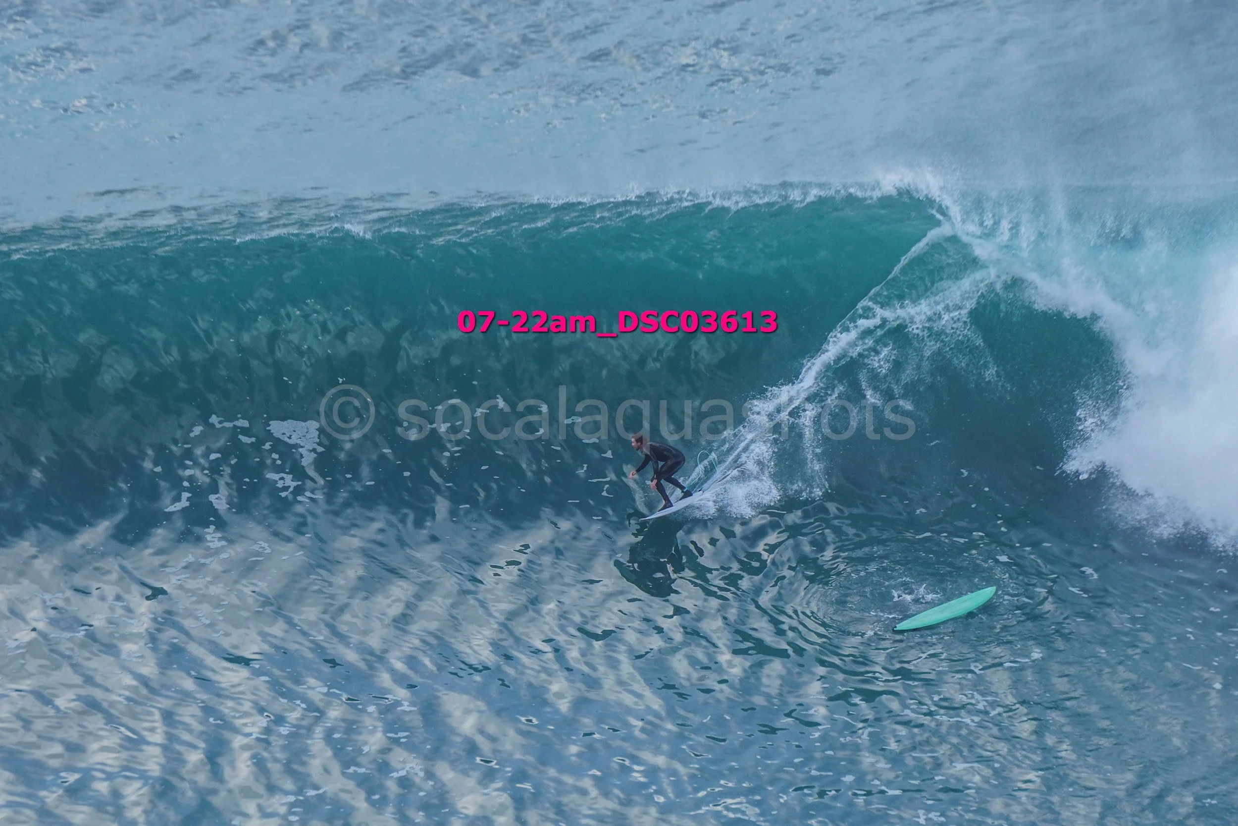 A person surfing on a large ocean wave with a light blue surfboard, wearing a wetsuit, captured at 7:22 am.