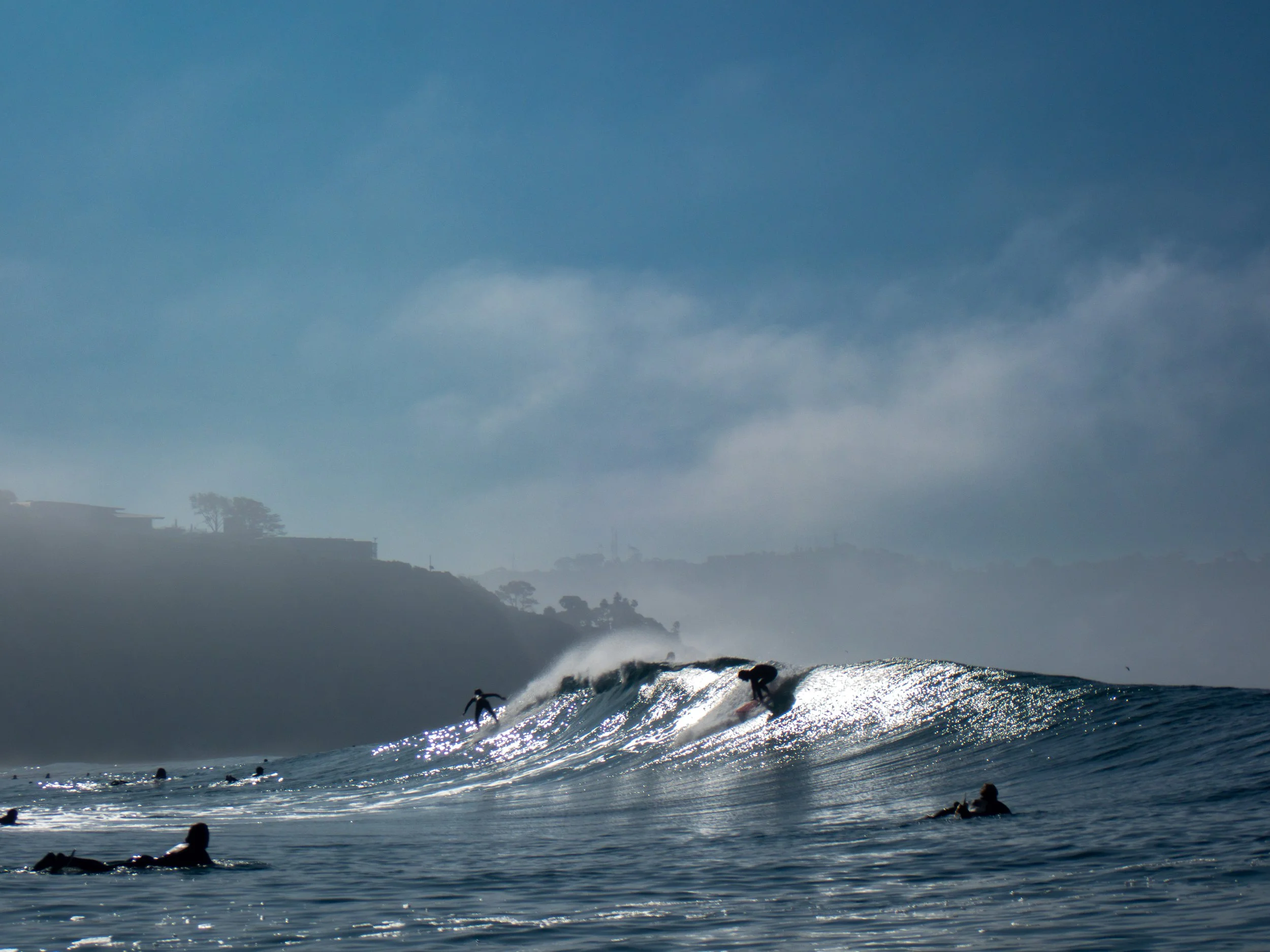 Surfers riding waves in the ocean with a misty coastline in the background under a blue sky.