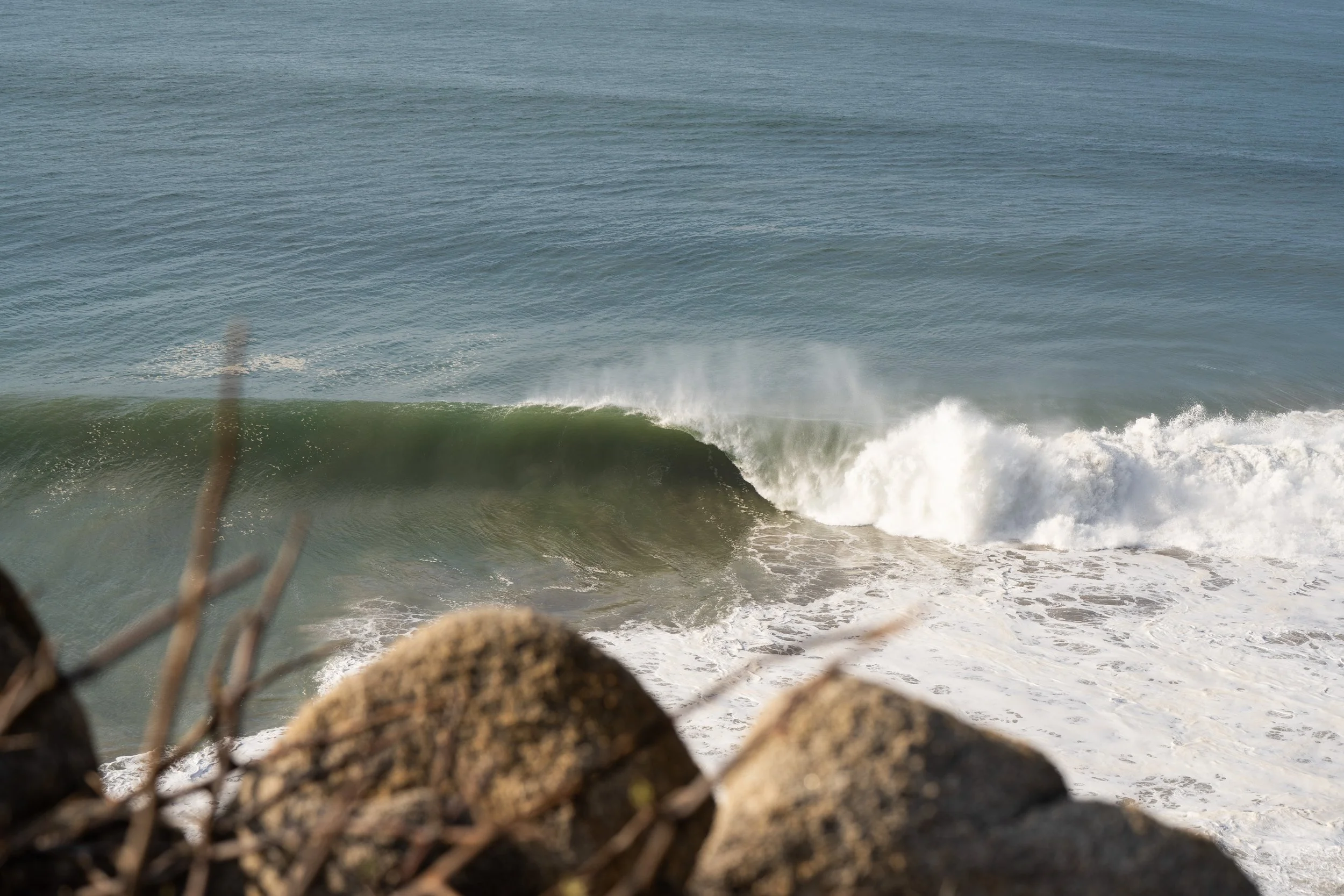 View of ocean waves crashing onto rocky shoreline, seen through a chain-link fence with some rocks in the foreground.