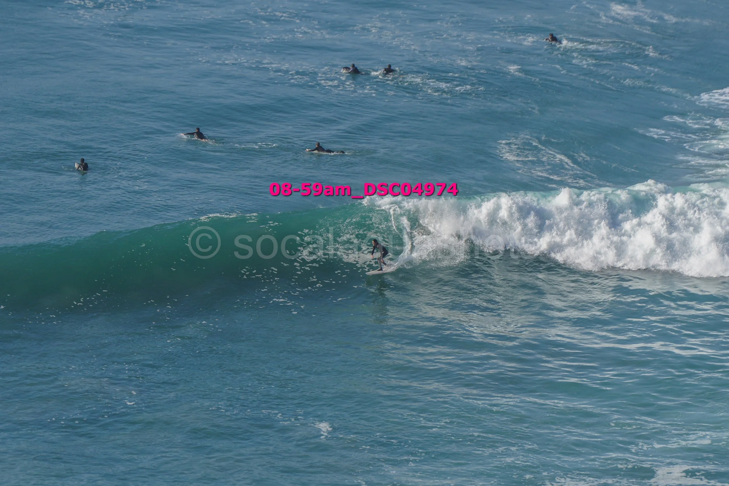 Surfer riding a wave in the ocean with several other surfers paddling in the water nearby.