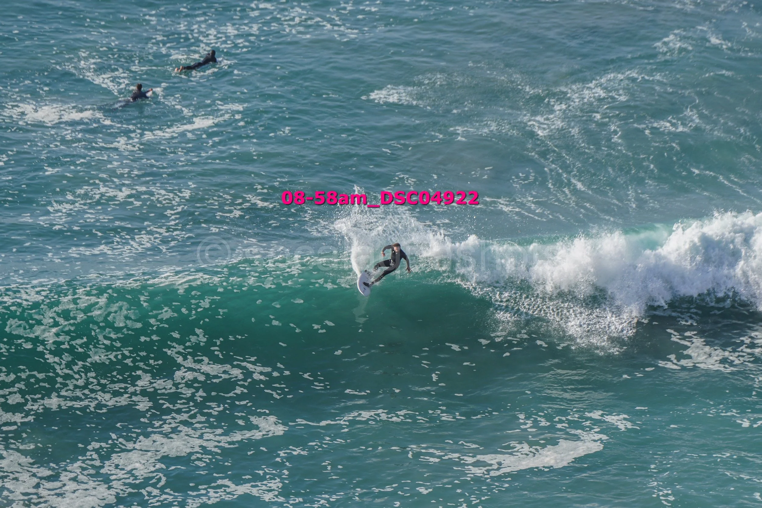 Surfer riding a wave in the ocean, with two other surfers paddling in the background.