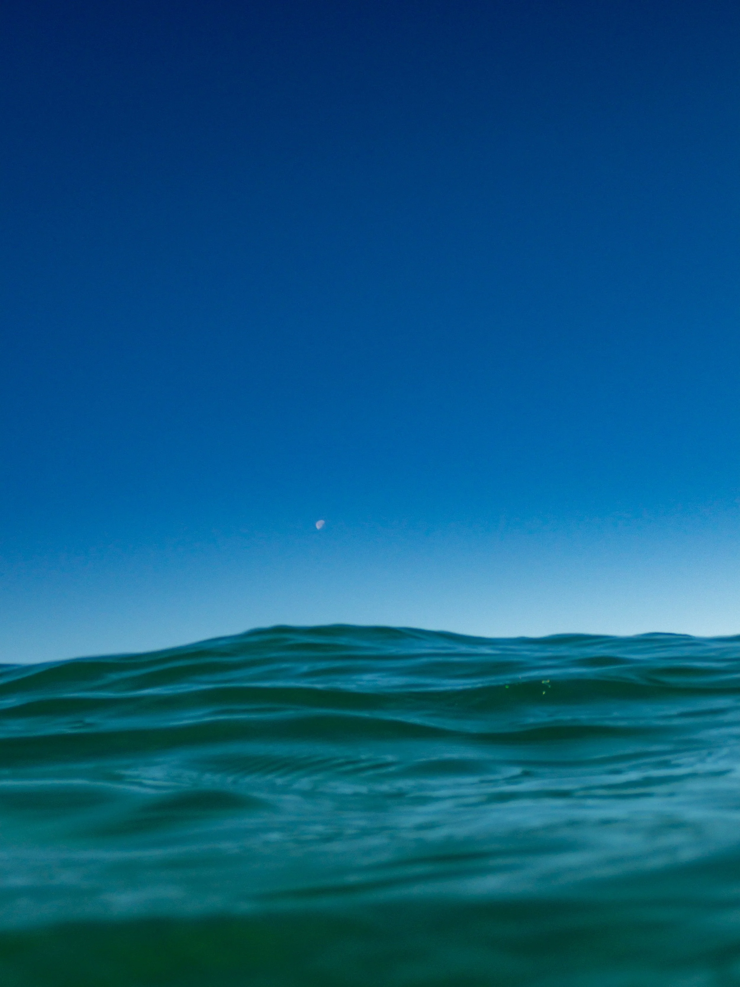 Ocean waves under a clear blue sky with the moon visible.