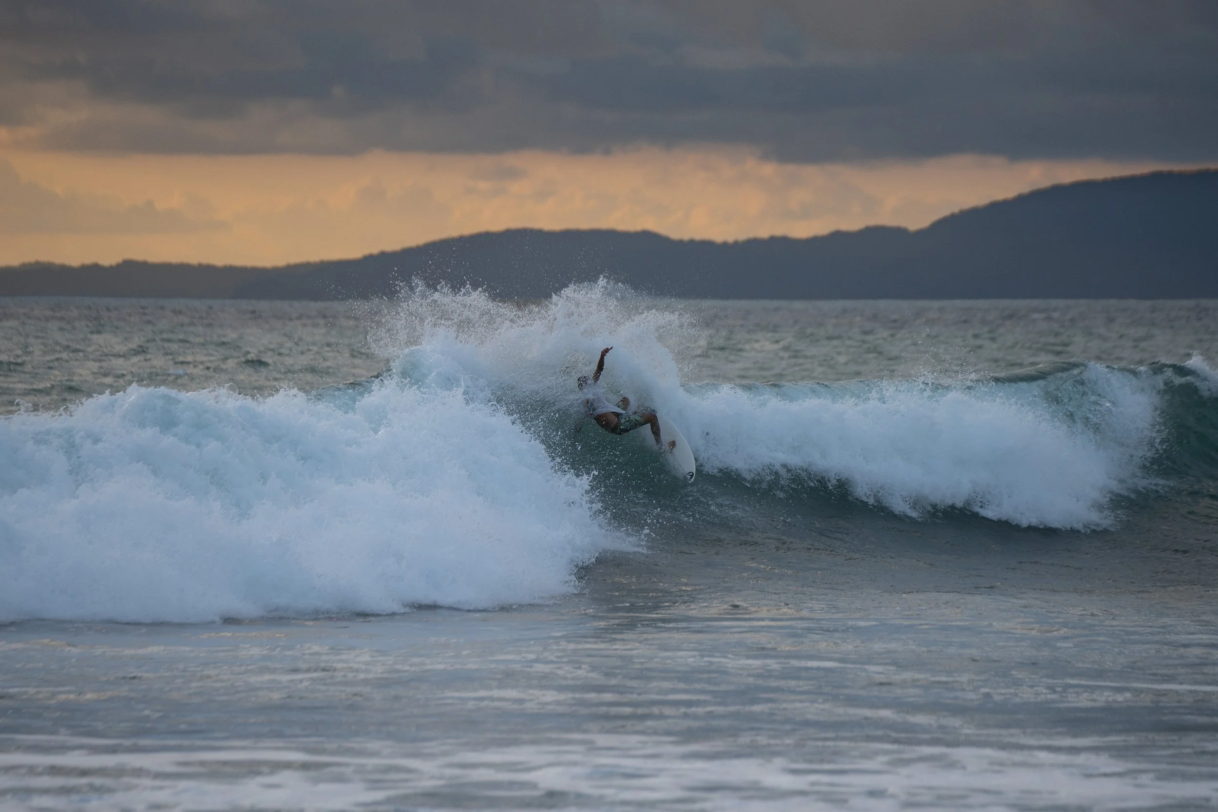 A person surfing on a wave in the ocean during sunset with mountains in the background.