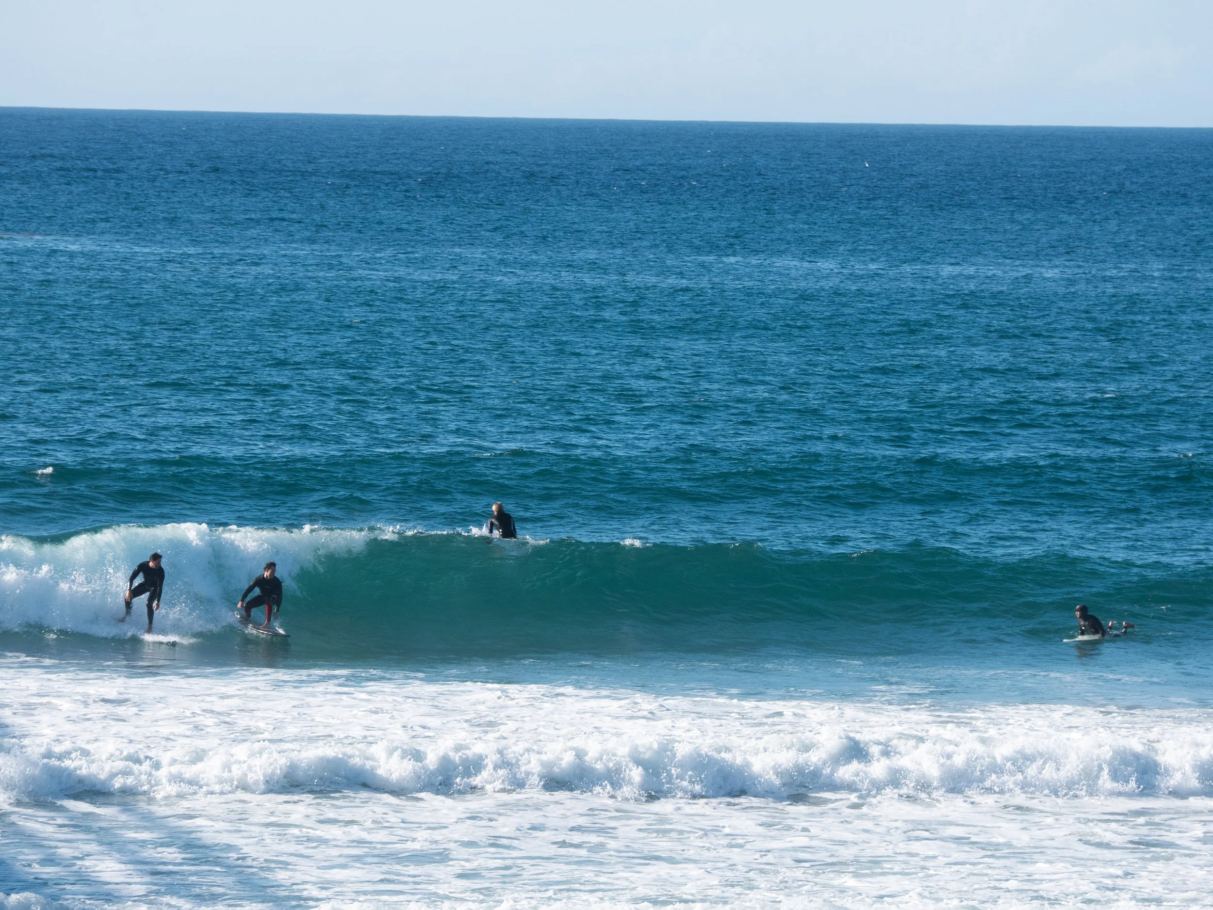 Four surfers in wetsuits riding and waiting for waves in the ocean with a clear blue sky.