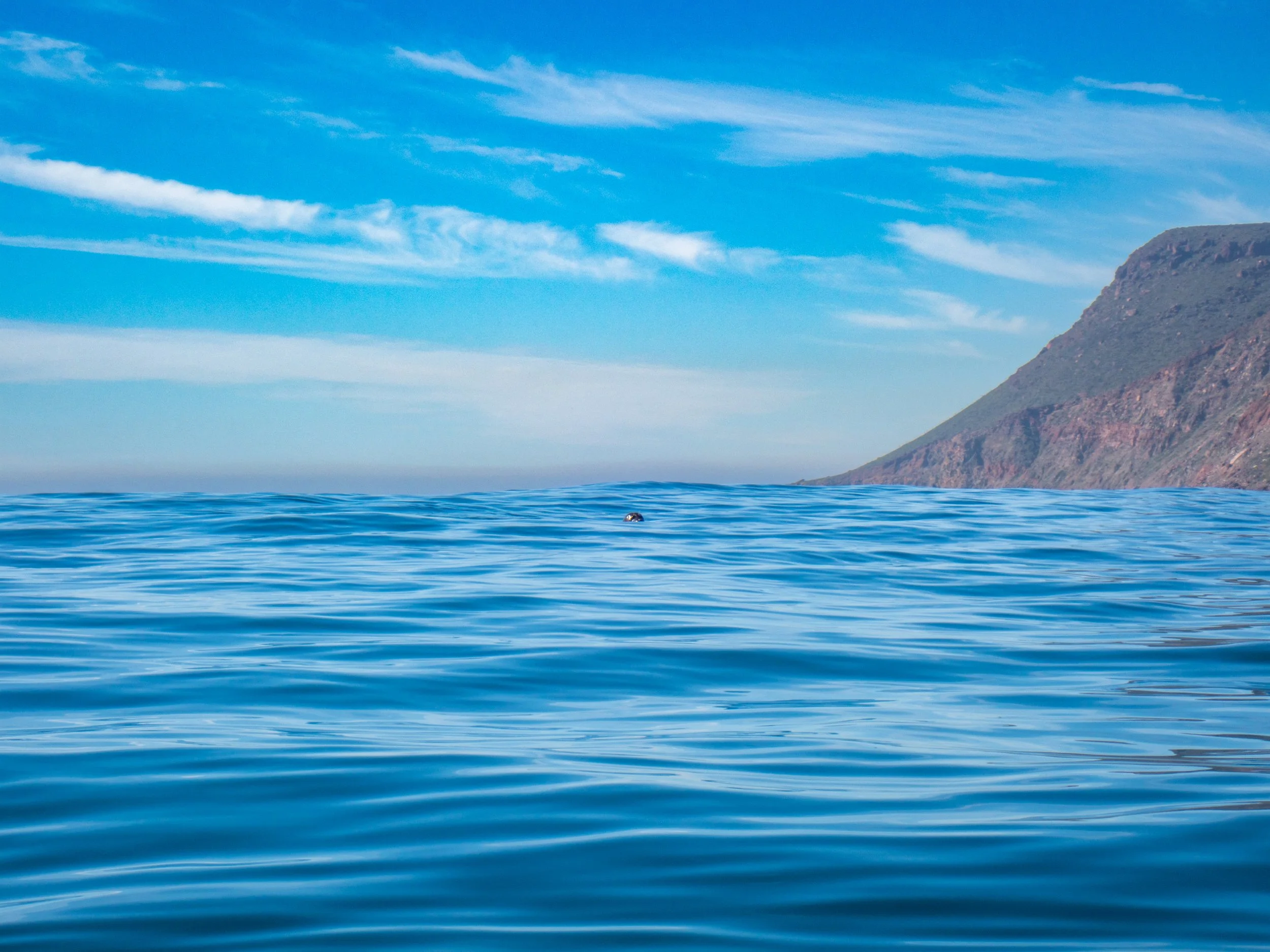 Ocean water with gentle ripples, mountain on the right side, partly cloudy blue sky.