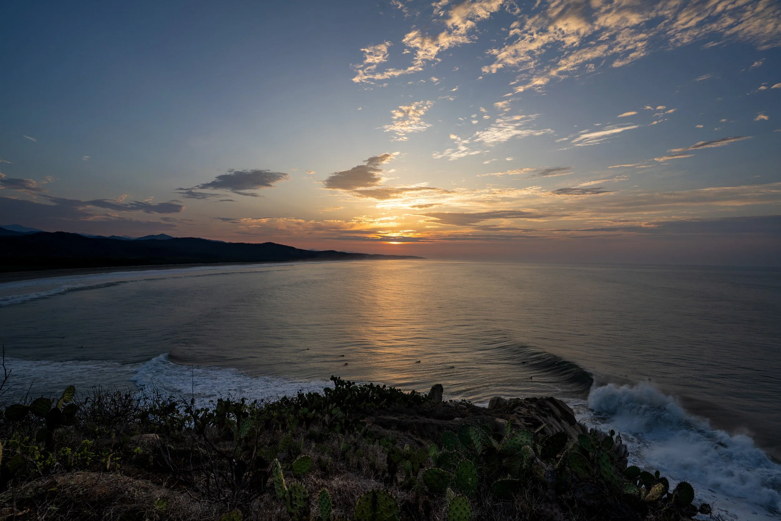 Sunset over the ocean with scattered clouds and silhouetted hills in the distance, rocky shoreline with cacti in the foreground.