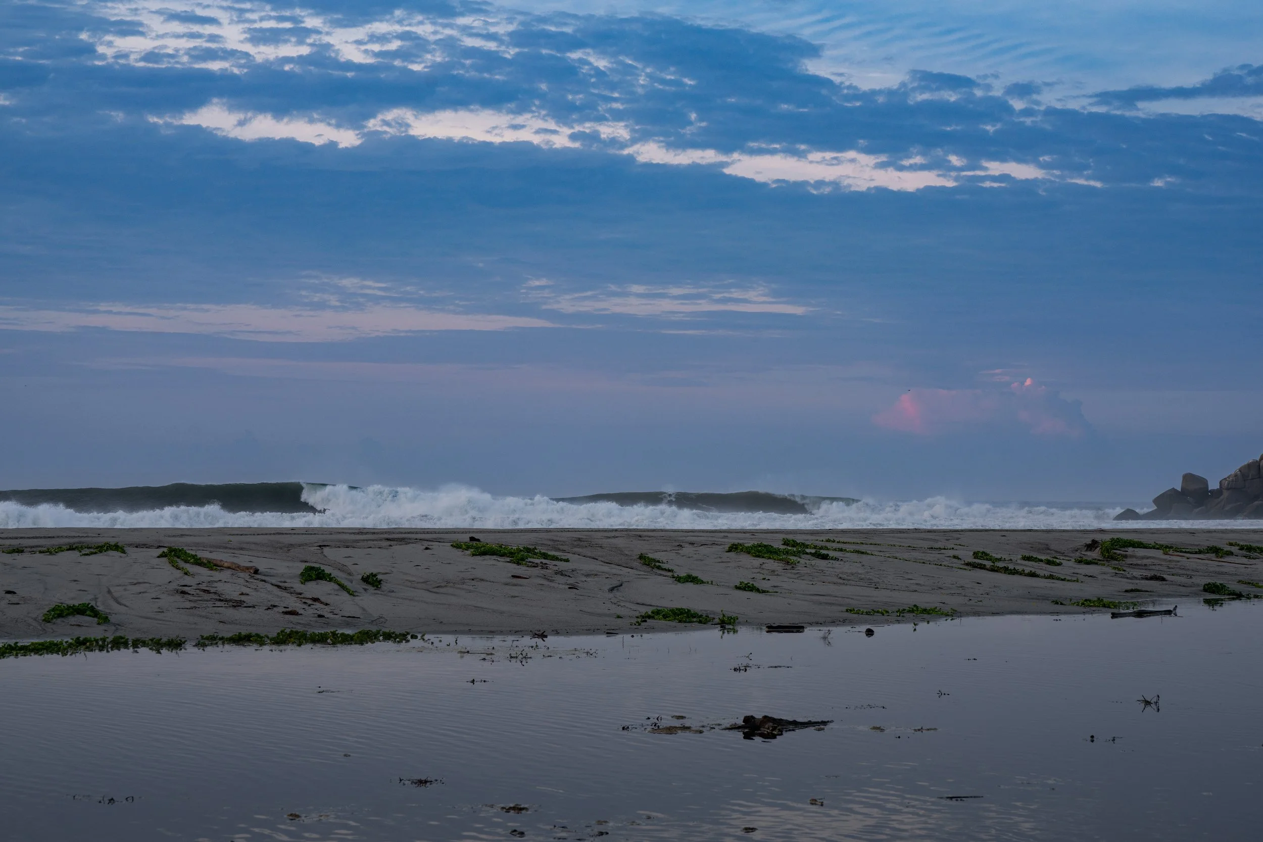 A landscape of a beach with a sandy shoreline, green vegetation, and ocean waves under a partly cloudy sky during dusk or dawn.