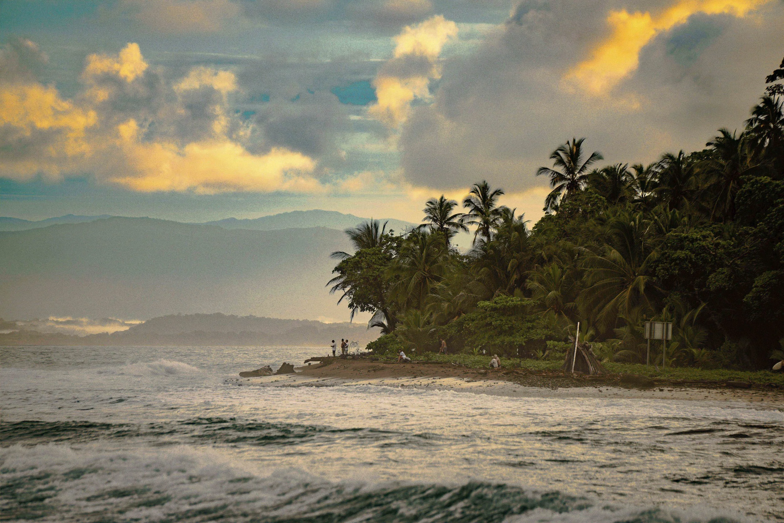 Beach scene with a shoreline, lush tropical trees on a hillside, and people relaxing on the sand, with mountains and cloudy sky in the background.