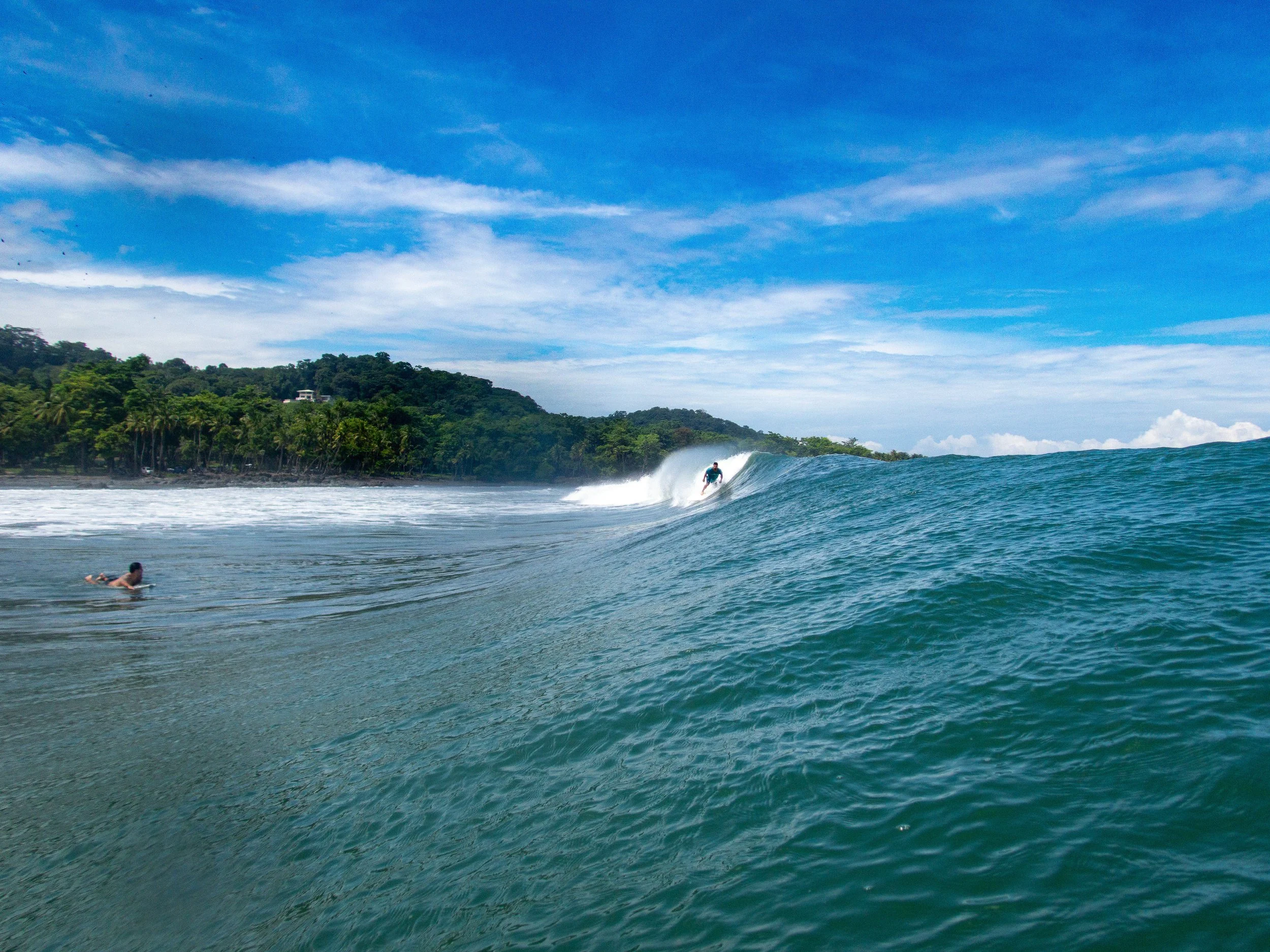 Surfer riding a large wave with another person on a surfboard in the ocean, tropical coastline in the background, under a blue sky.