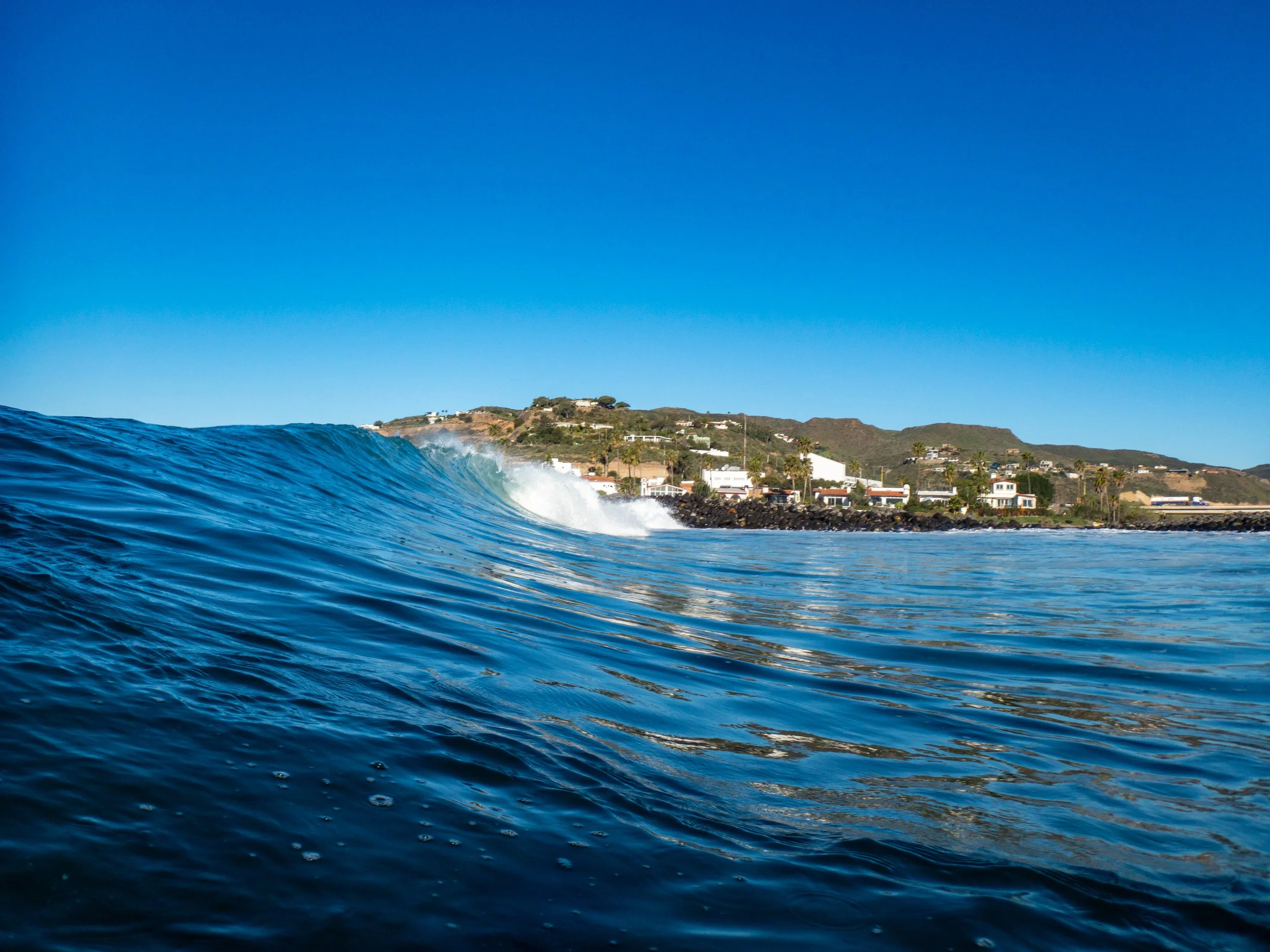Ocean wave with a coastal town and green hills in the background under a clear blue sky.