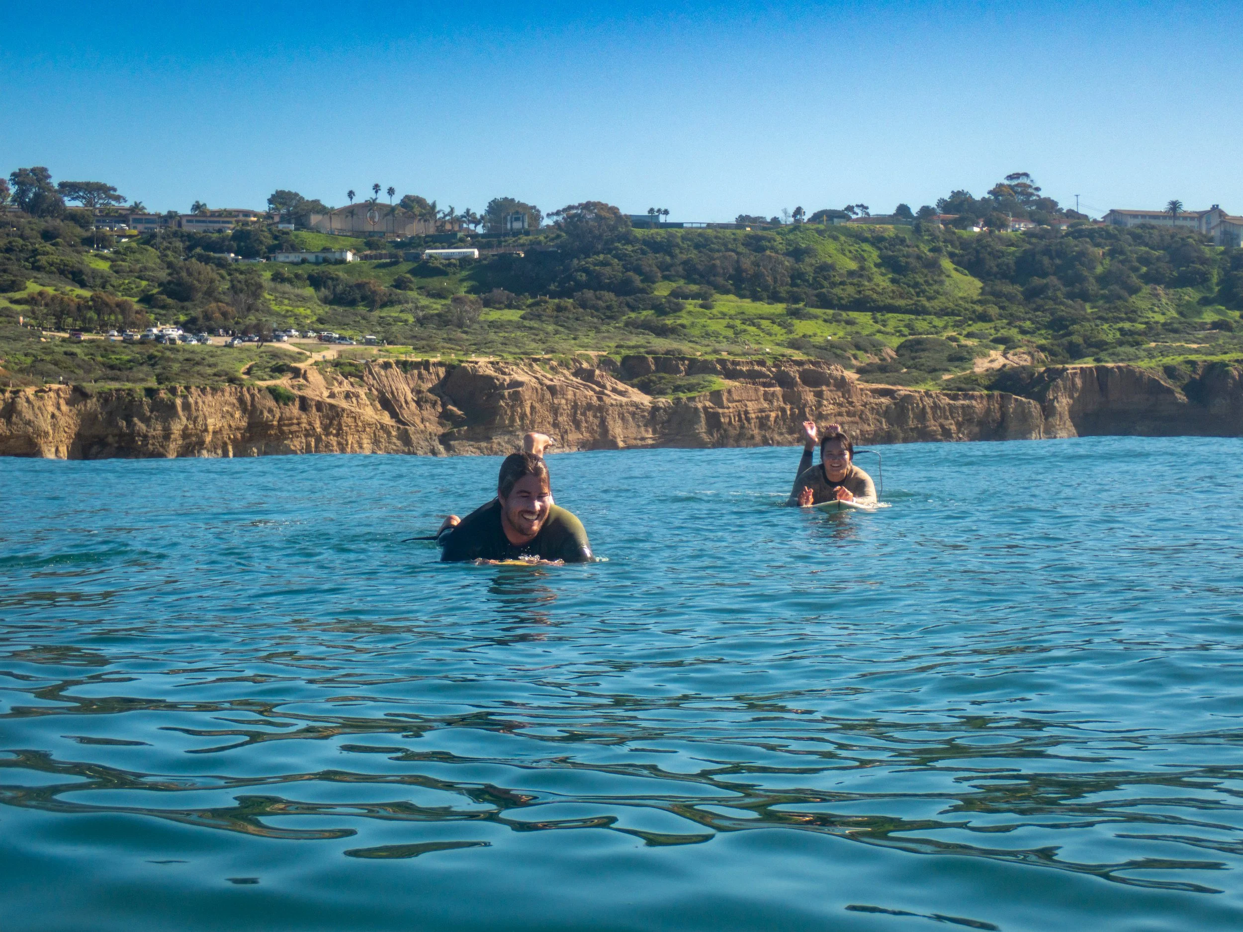 Two people swimming in the ocean with a rocky coastline and green hills in the background on a sunny day.