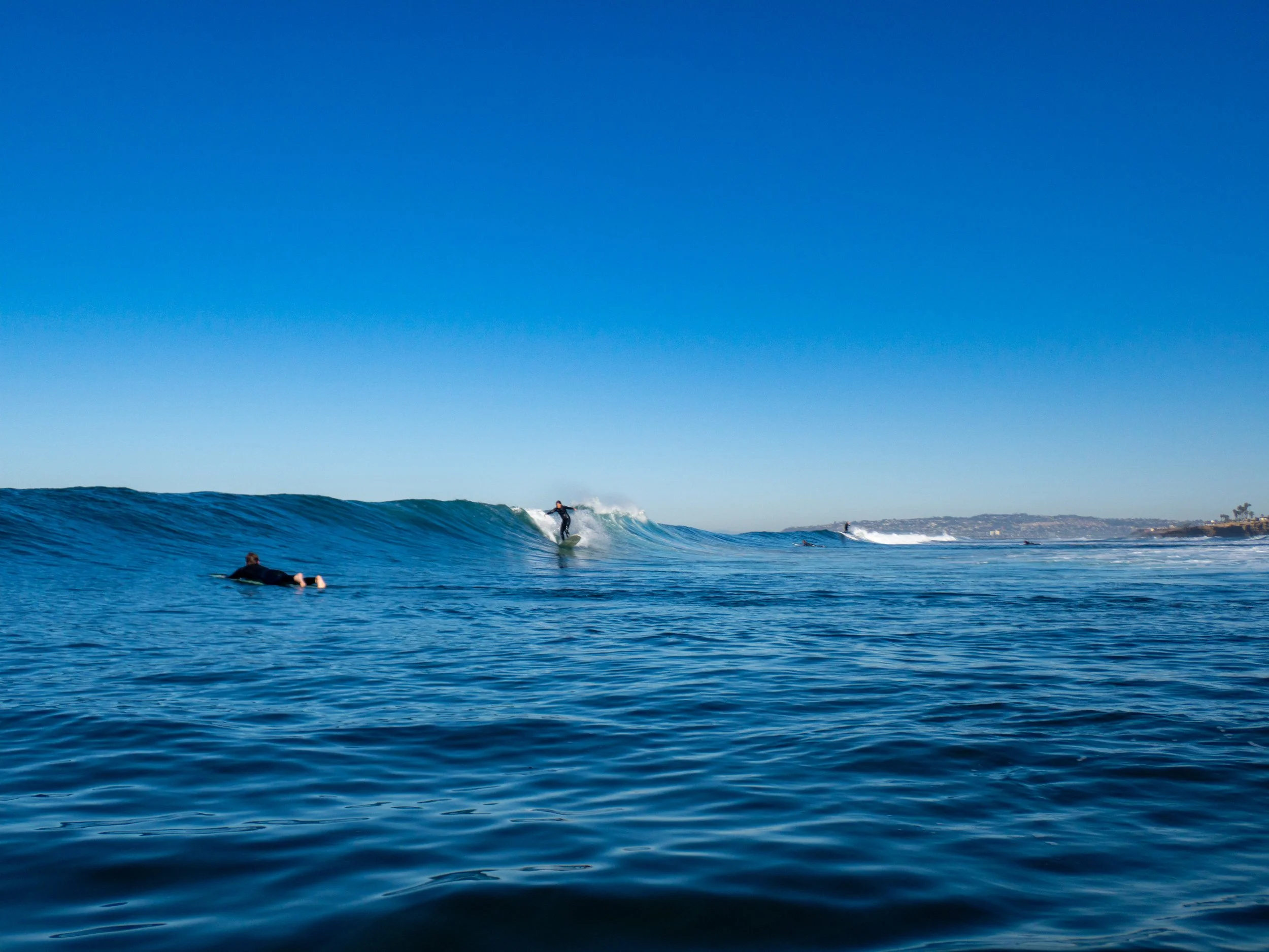 A person surfing on a wave in the ocean with a clear blue sky, and another person floating nearby