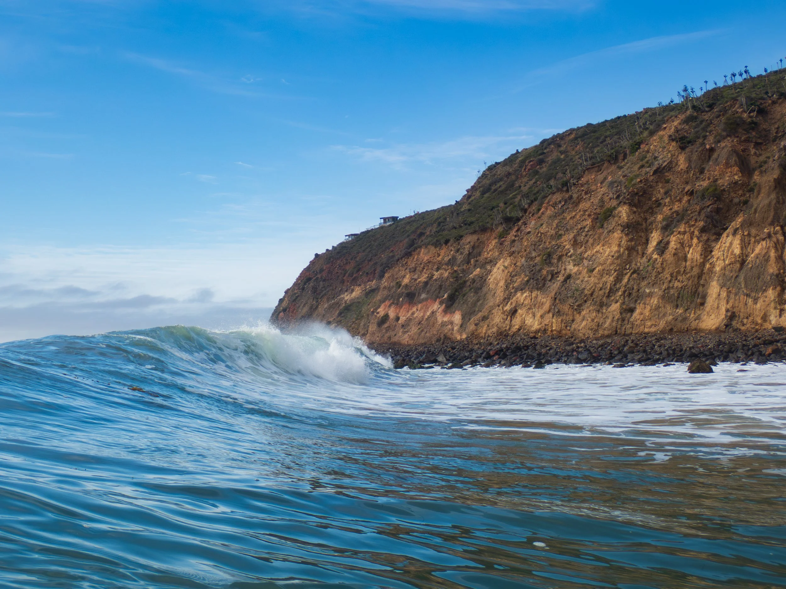 Ocean waves crashing near a rocky coastline with a steep hillside in the background, under a blue sky with scattered clouds.