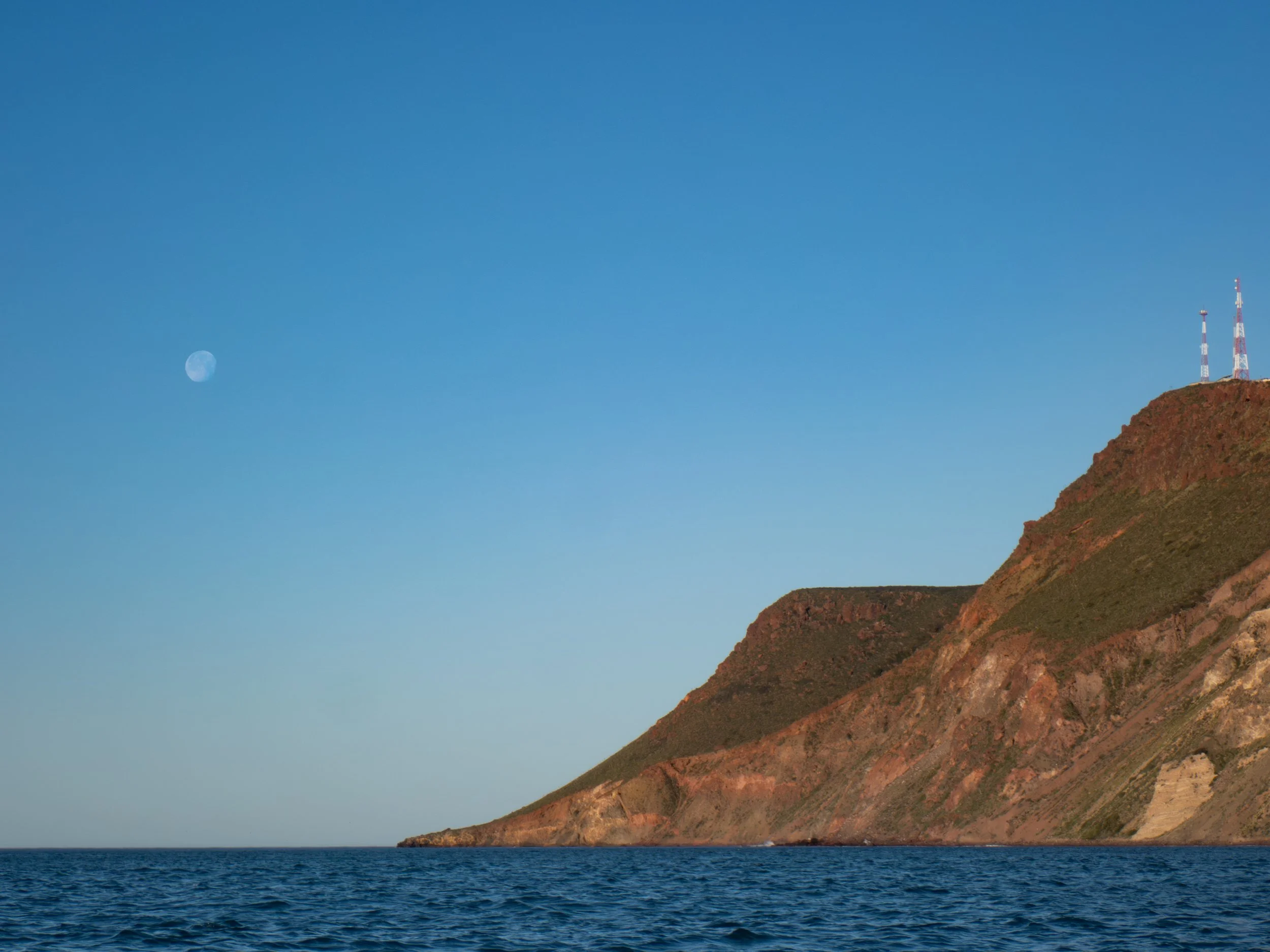 A view of a rocky, reddish-brown hillside with antennas on top, against a clear blue sky with a visible moon and calm ocean water in the foreground.