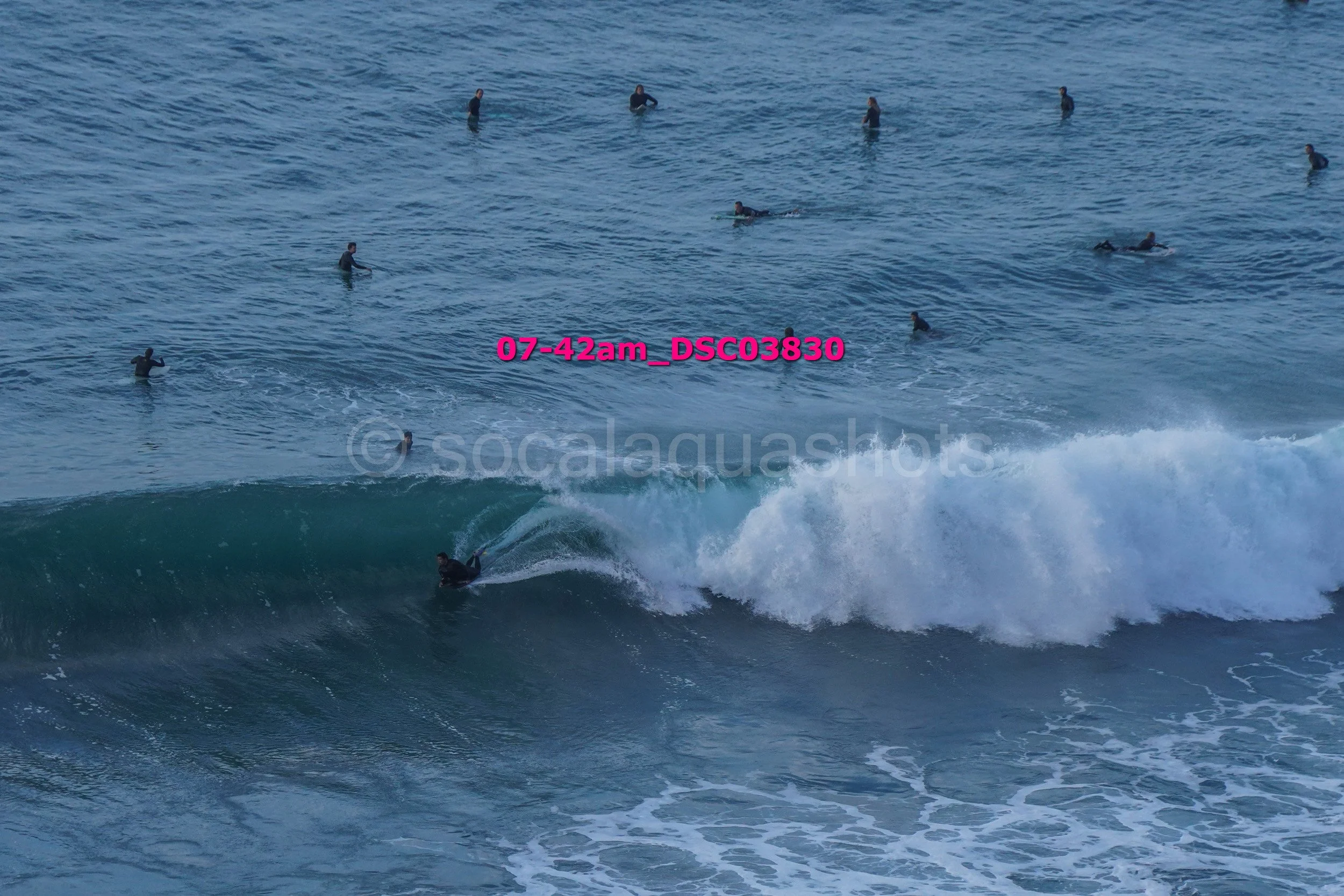 A surfer riding a wave in the ocean with several swimmers or surfers in the background, with the timestamp 07-42am and the watermark 'socalaquashots'.