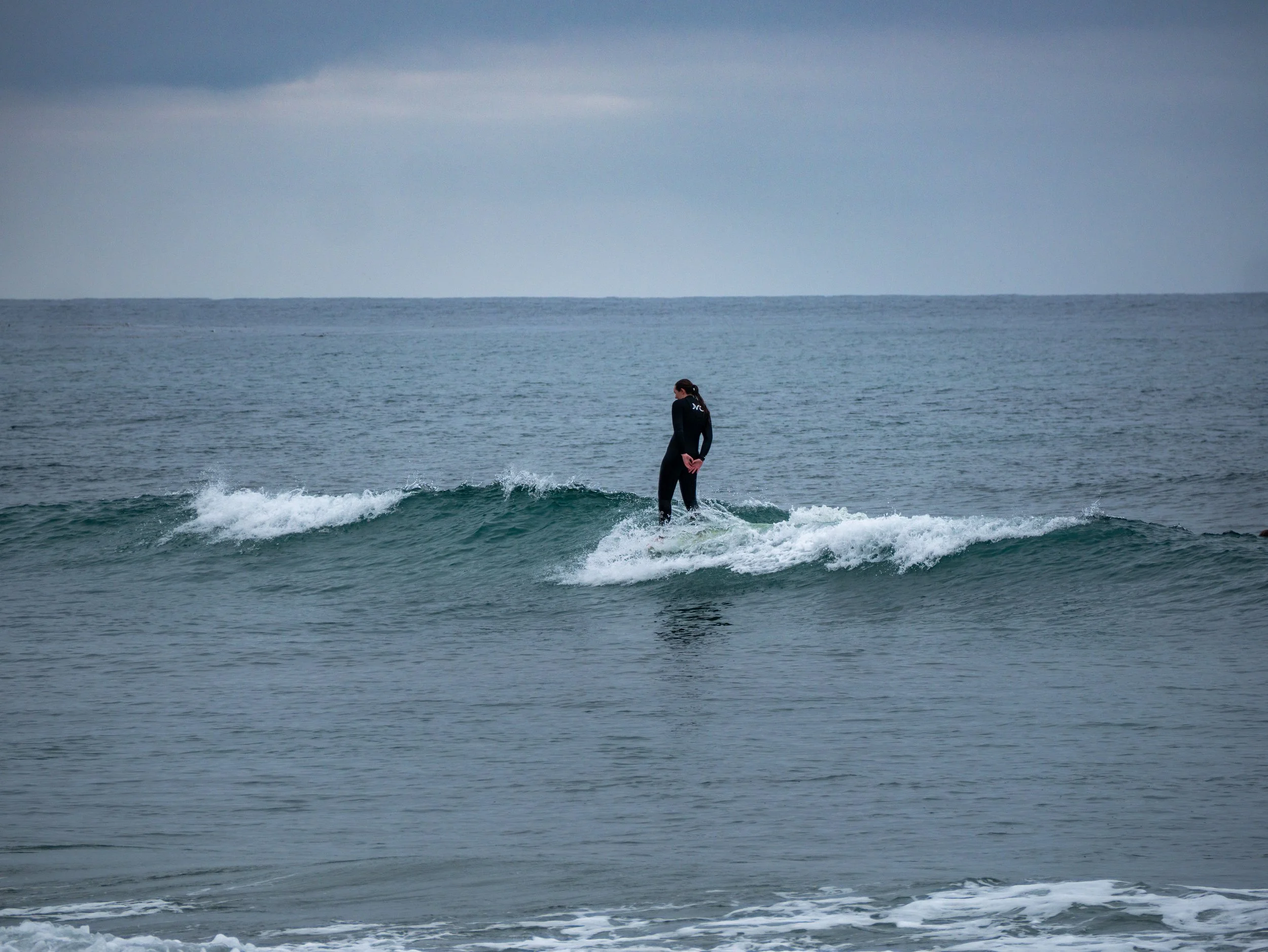 A person in a wetsuit standing on a surfboard riding a small wave in the ocean with a cloudy sky above.