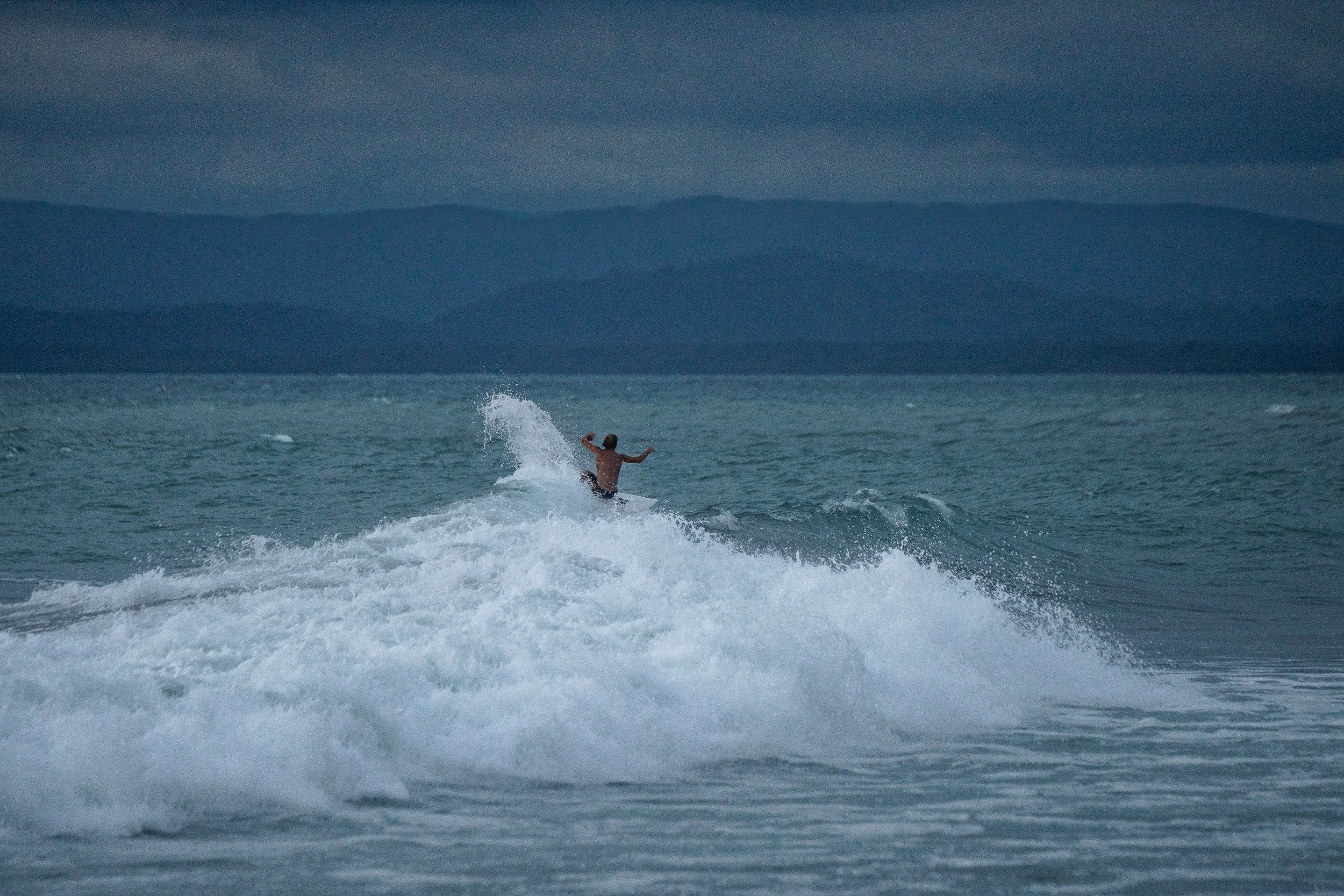 A person surfing on a wave in the ocean, with dark, cloudy skies and distant mountains in the background.