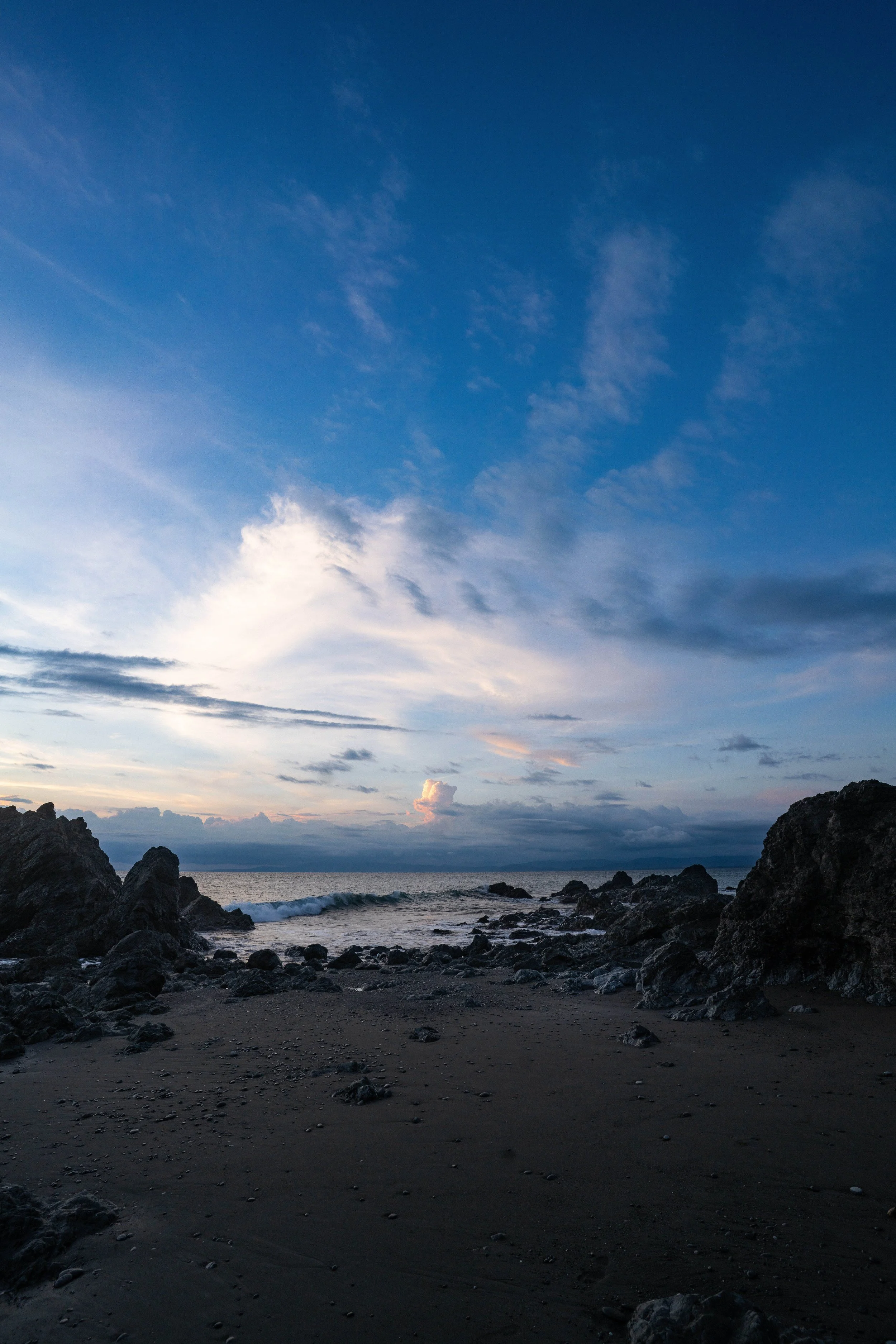 A rocky beach with dark sand, scattered small rocks and larger boulders. The ocean waves are gently hitting the shore. The sky is partly cloudy with blue and gray clouds, indicating either dawn or dusk.