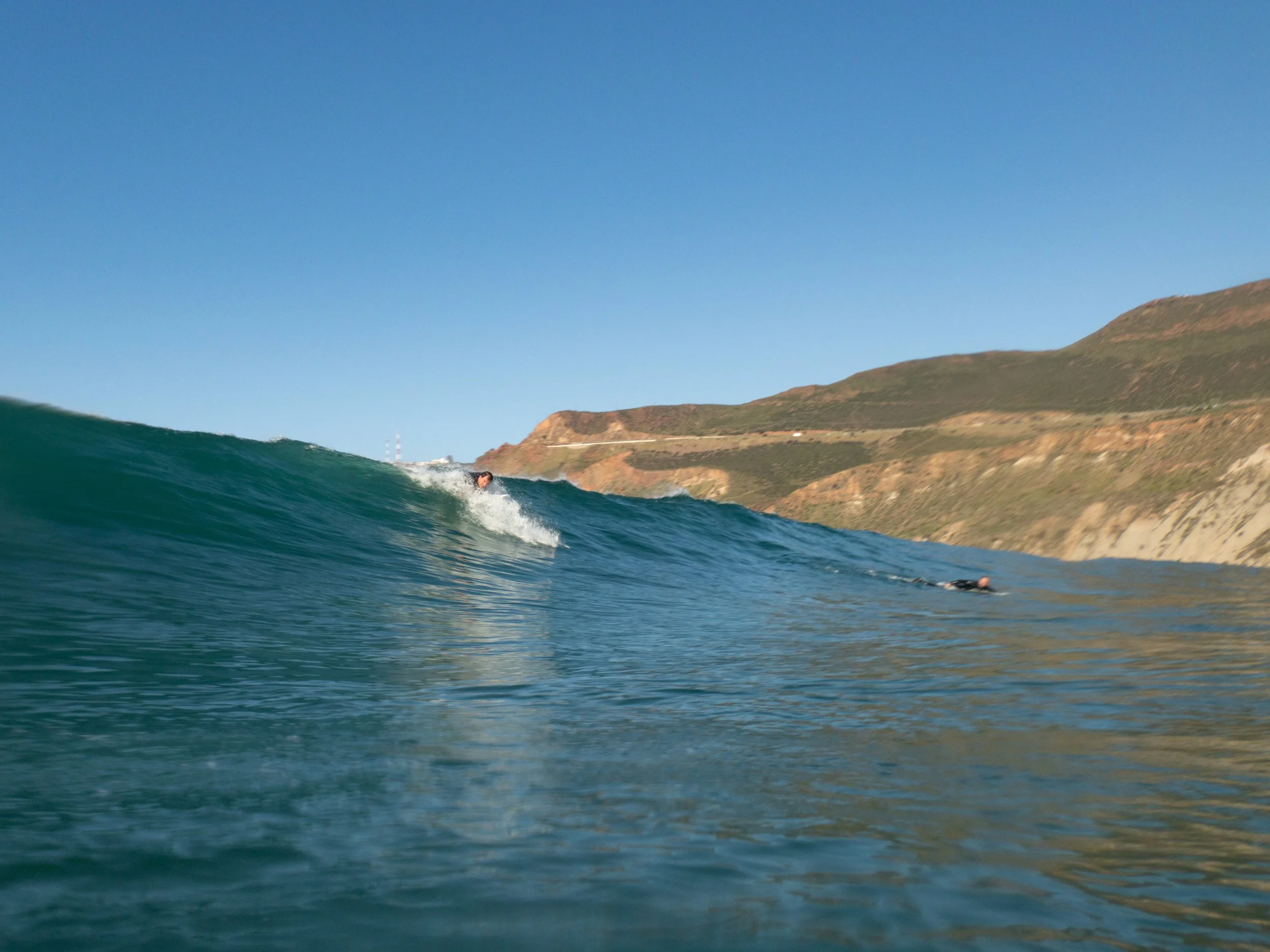 Two surfers riding a wave at the beach with hills in the background, clear blue sky, and calm water.