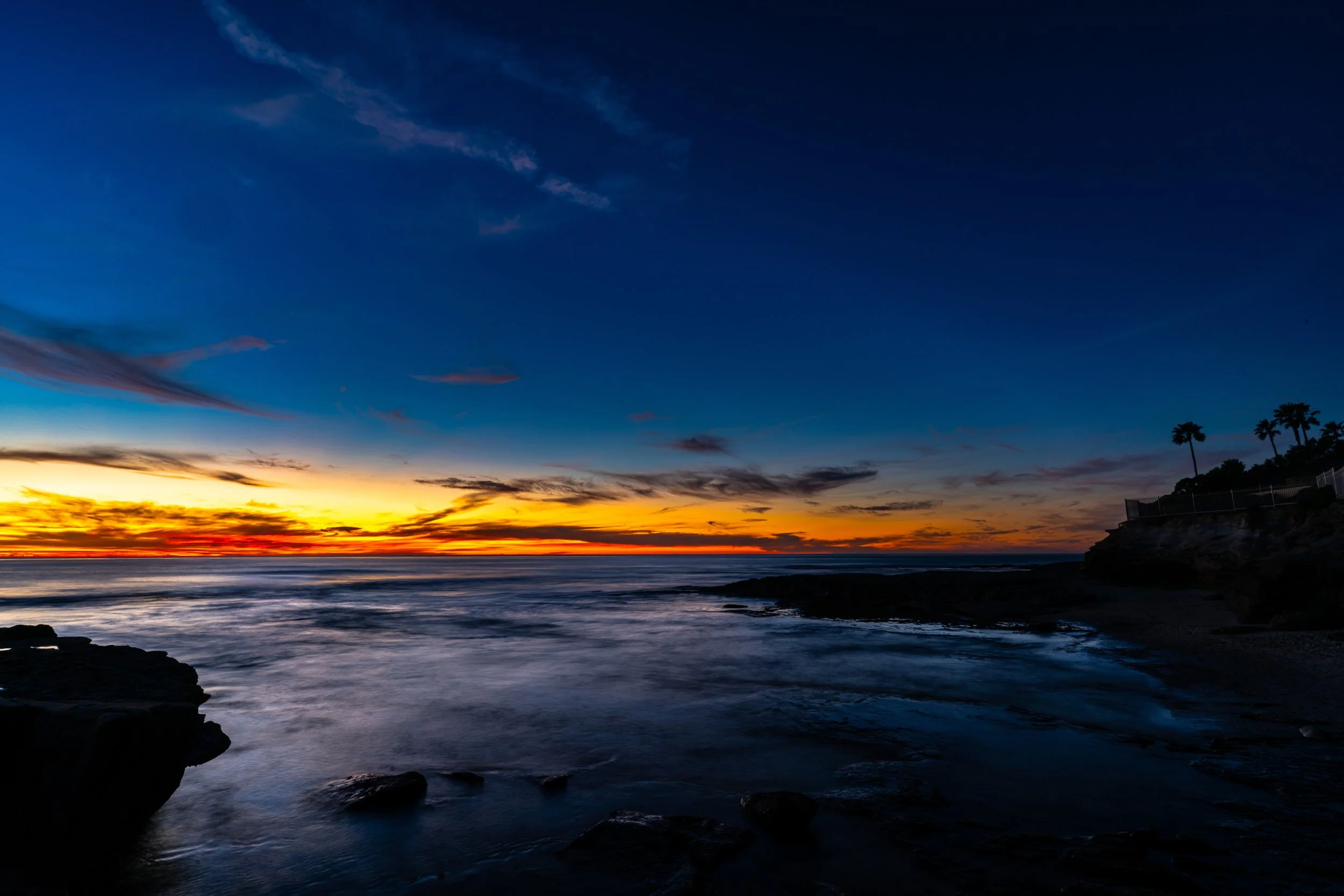 Sunset over the ocean with colorful sky and palm trees on a rocky coastline.