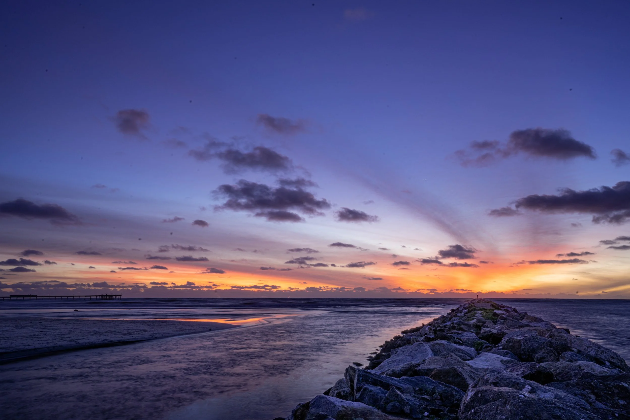 A sunset over the ocean with a rocky jetty extending into the water, clouds in the sky, and a pier in the background.