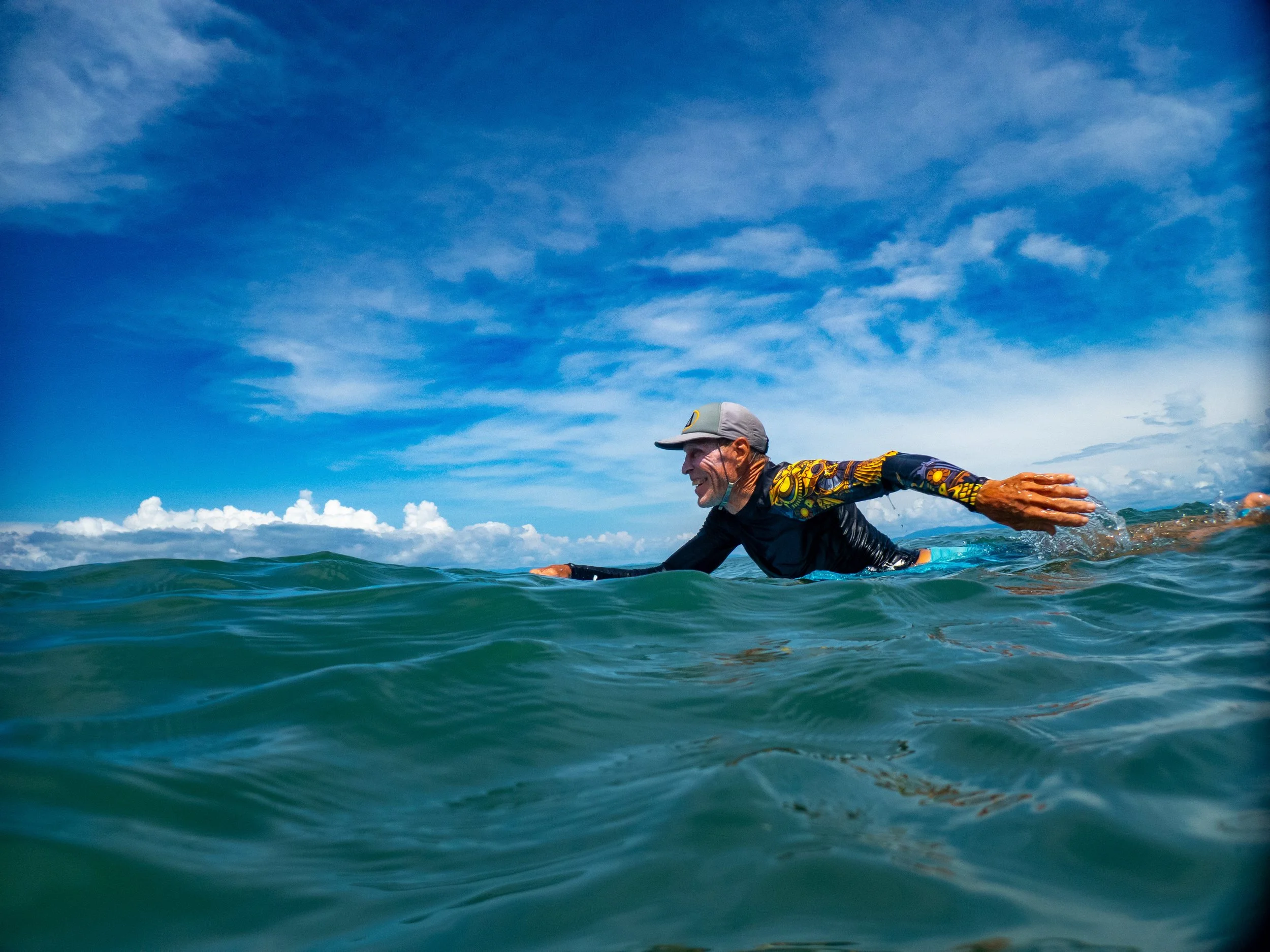 An elderly man swimming in open water under a partly cloudy sky, wearing a cap and a long-sleeve wetsuit with a colorful design on the sleeves.