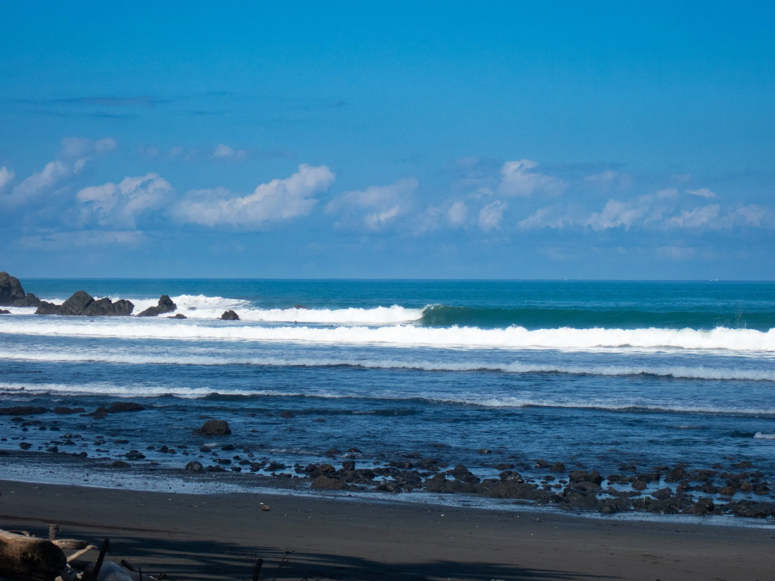 View of the ocean with waves, rocky shoreline in the foreground, and a partly cloudy sky.