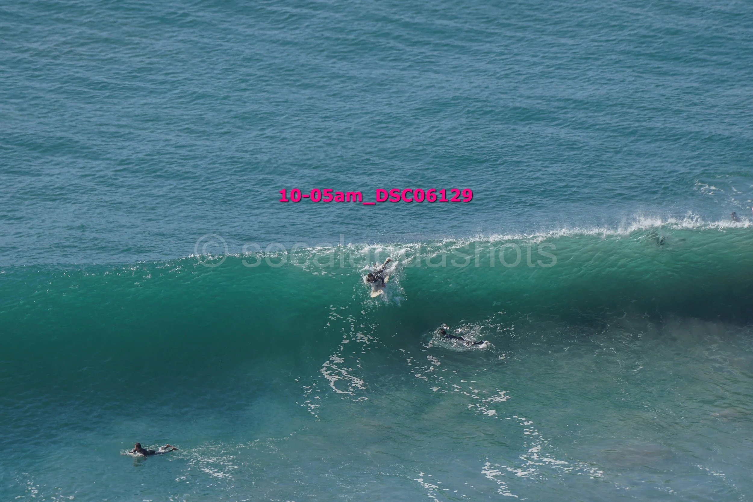 Surfers riding a large ocean wave at sunrise, with some surfers paddling on their boards in the water.