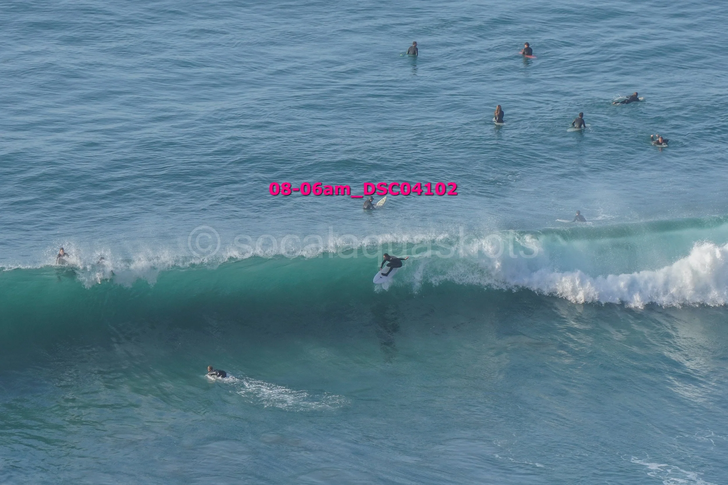 A person in a wetsuit riding a surfboard on a large wave with others swimming and surfing in the background in the ocean.
