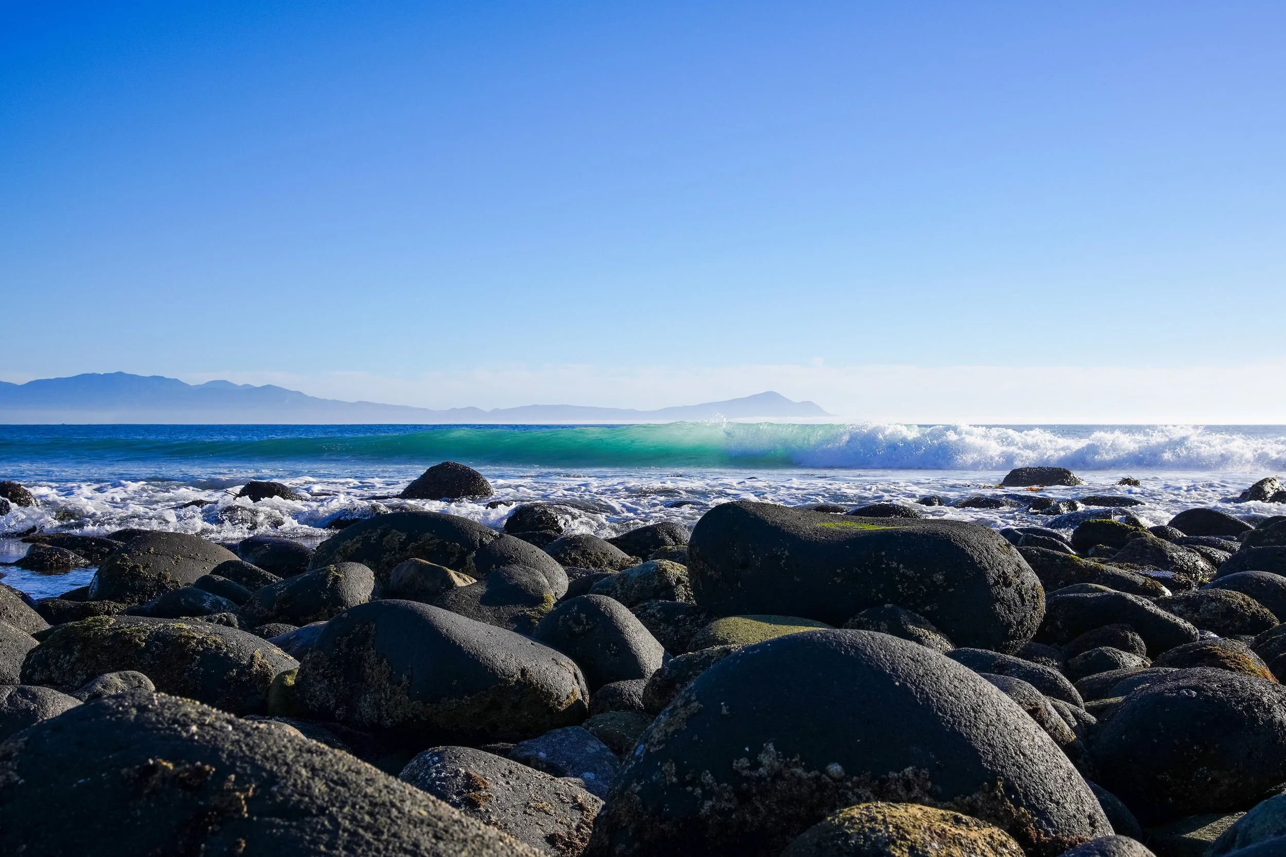 Large black rocks along a sunny beach with blue ocean and clear sky, distant mountains in the background.