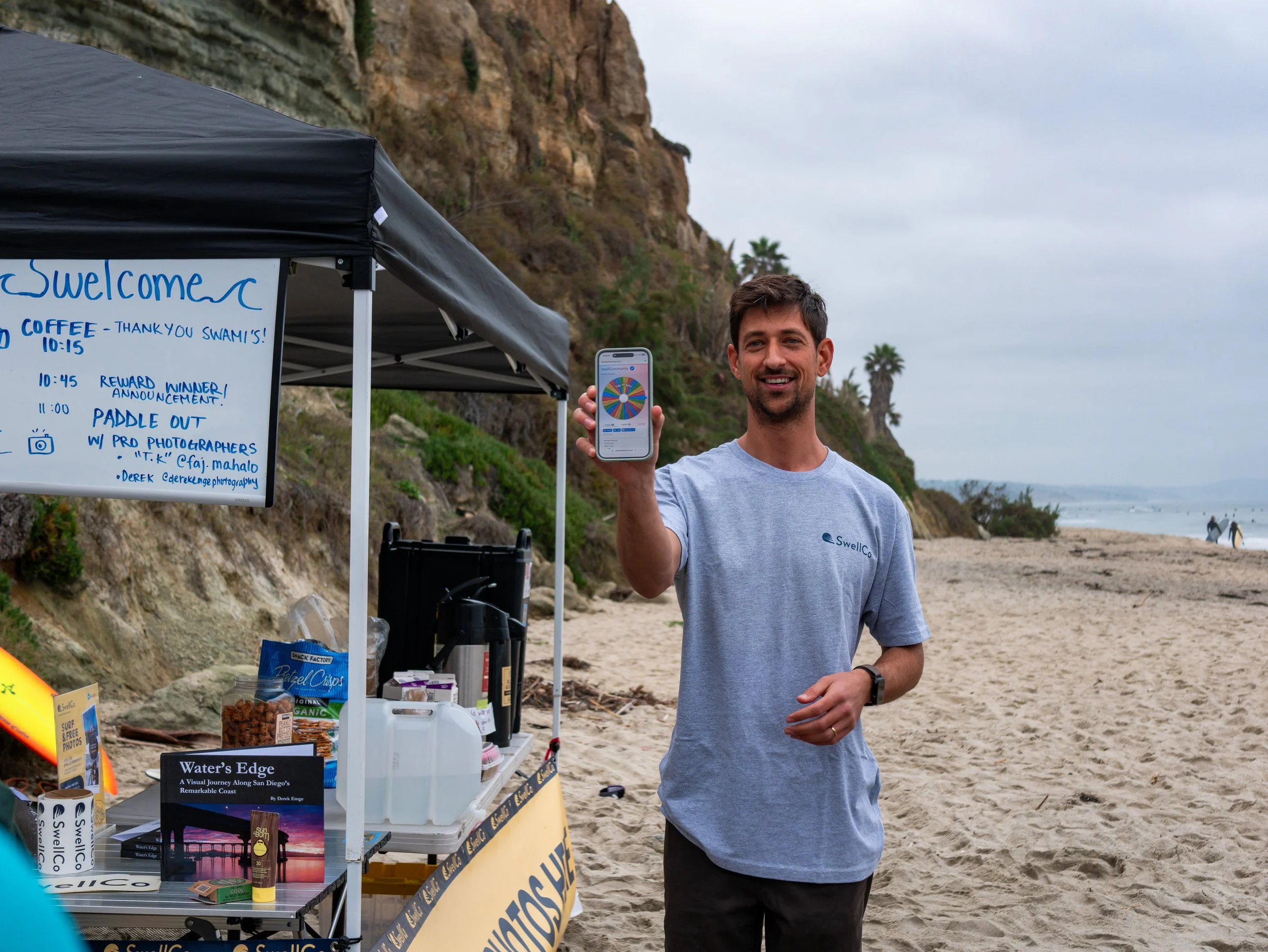 A man smiling on a beach holding a smartphone displaying a colorful wheel or game. Behind him is a beachside stand with a whiteboard detailing schedule and snacks, and a scenic coastline with cliffs and distant people walking on the sand.