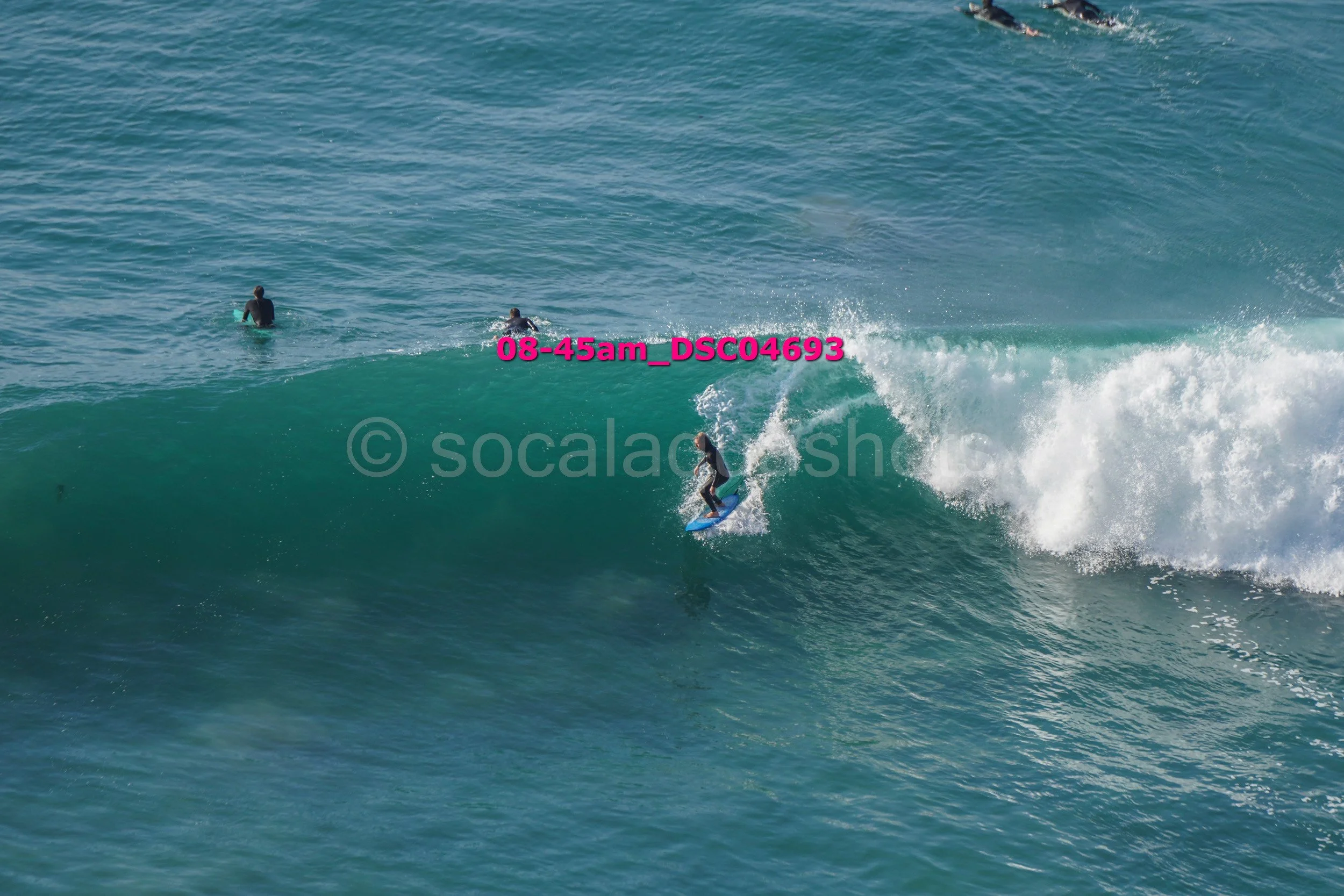 A person surfing on a blue surfboard on a large wave in the ocean with two other surfers in the background.