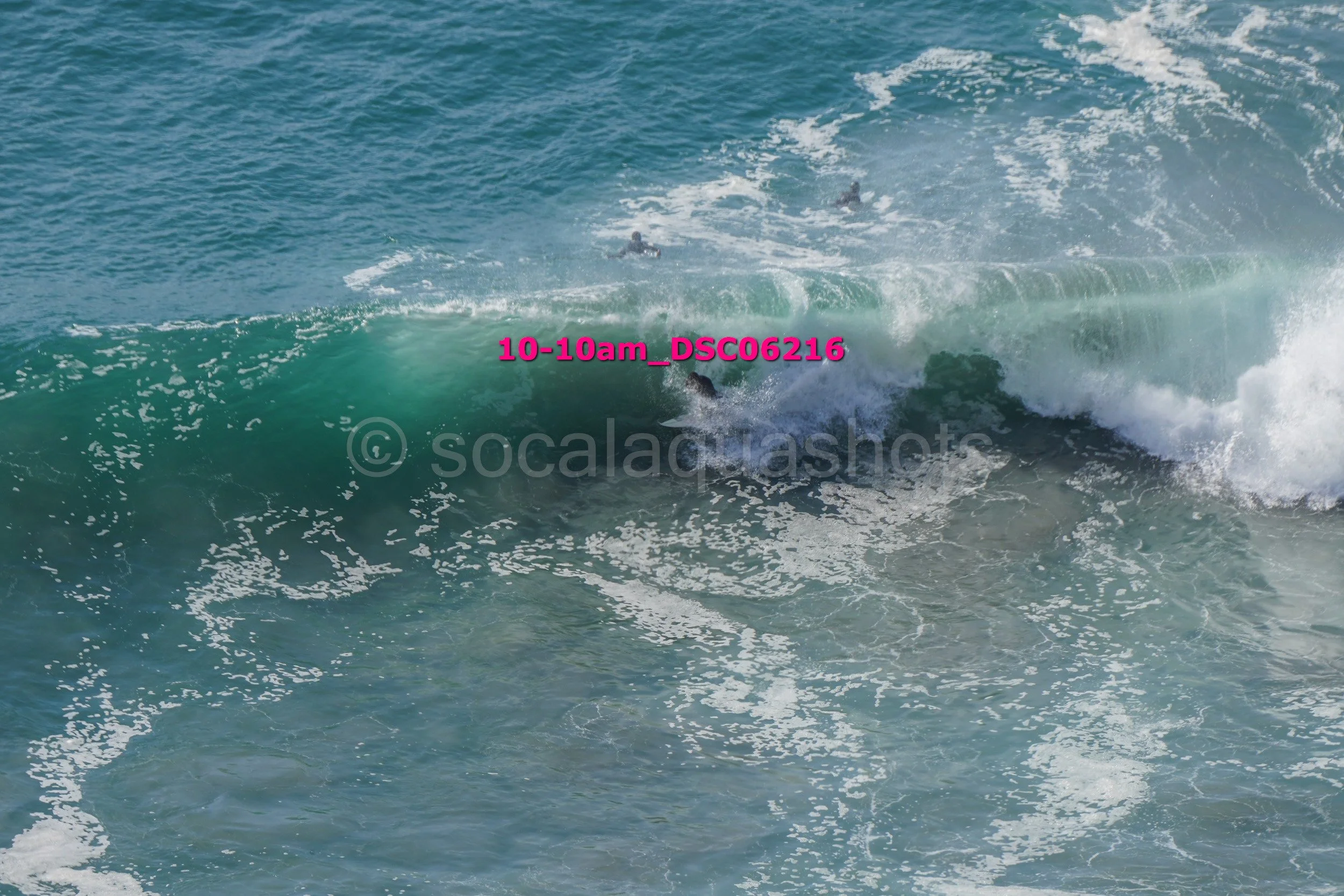 Surfers riding a large ocean wave in the water, with some surfers visible in the background.