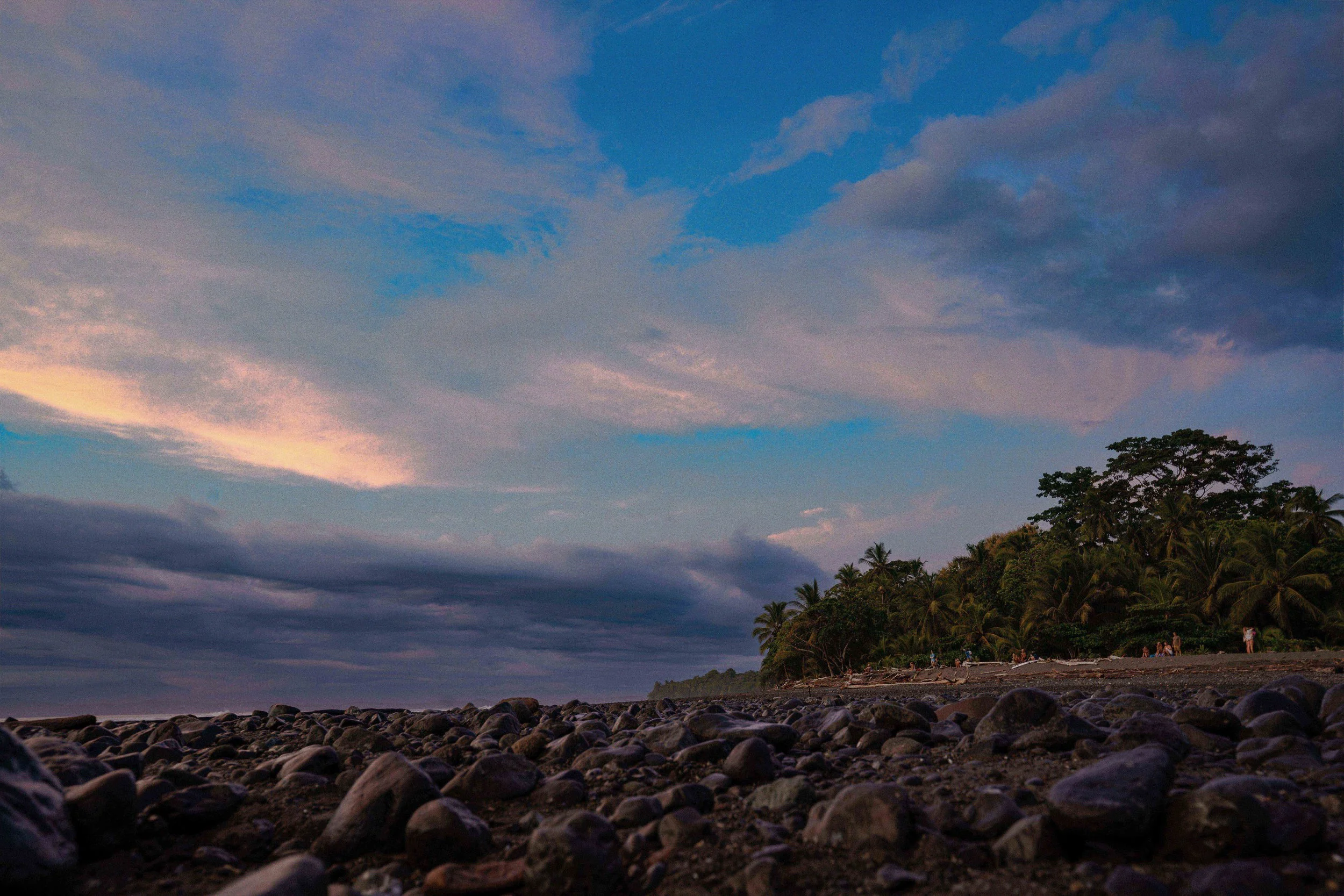 A rocky beach at sunset with a view of a distant horizon and a partly cloudy sky with blue and purple hues. There are trees along the shoreline and some people walking or standing near the trees.
