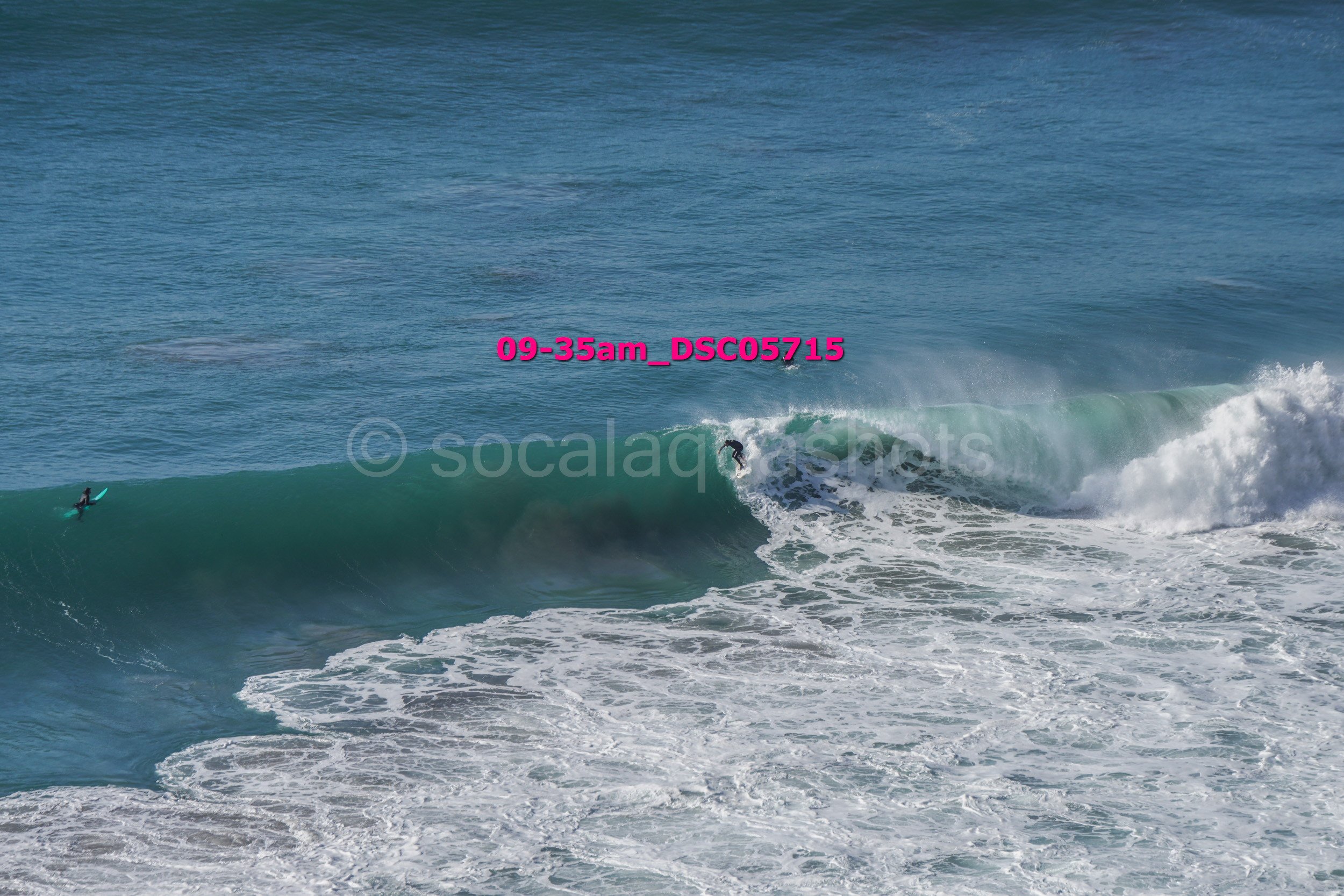 Two surfers riding a large wave in the ocean, with one surfer on the wave and the other waiting on a surfboard nearby.